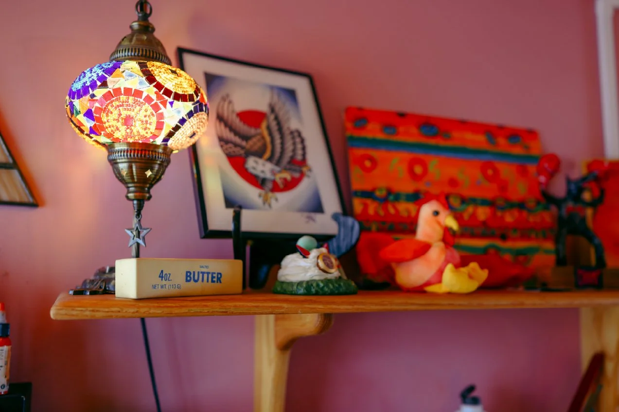 Decorative room with a colorful mosaic lamp, framed artwork of an eagle, a small duck figurine, and plush duck toys on a wooden shelf against a pink wall.