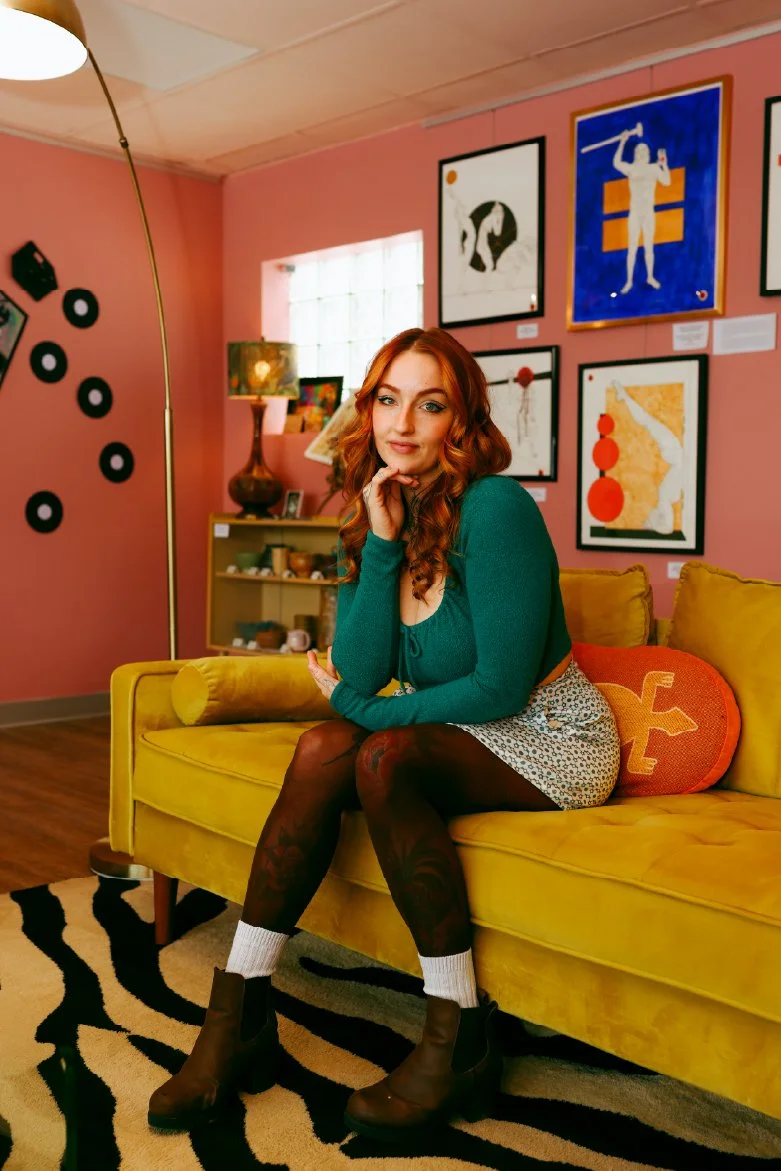 A woman with red hair sitting on a yellow couch in a colorful living room with pink walls, framed artwork, and a black and white patterned rug.