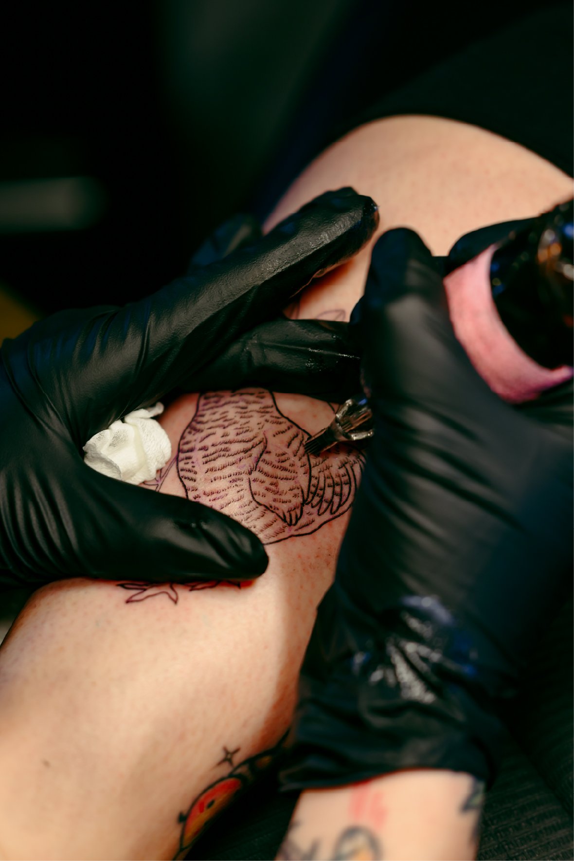 A person getting a tattoo of a line drawing of an eagle on their leg, with a tattoo artist using a tattoo machine and wearing black gloves.
