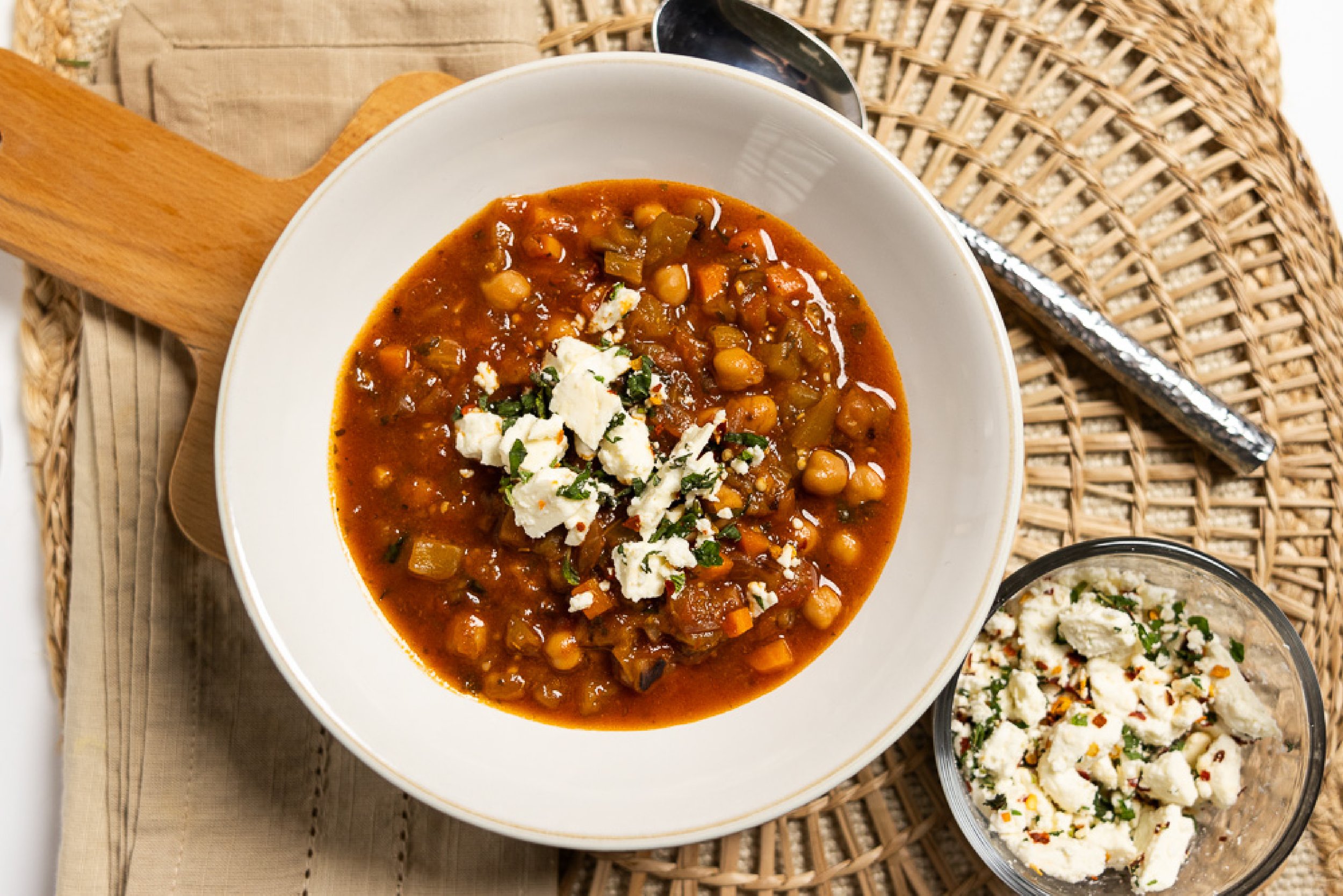 A bowl of chickpea and vegetable stew topped with cheese and herbs, placed on a wooden cutting board. Nearby, a smaller bowl contains a creamy potato salad with herbs and spices.