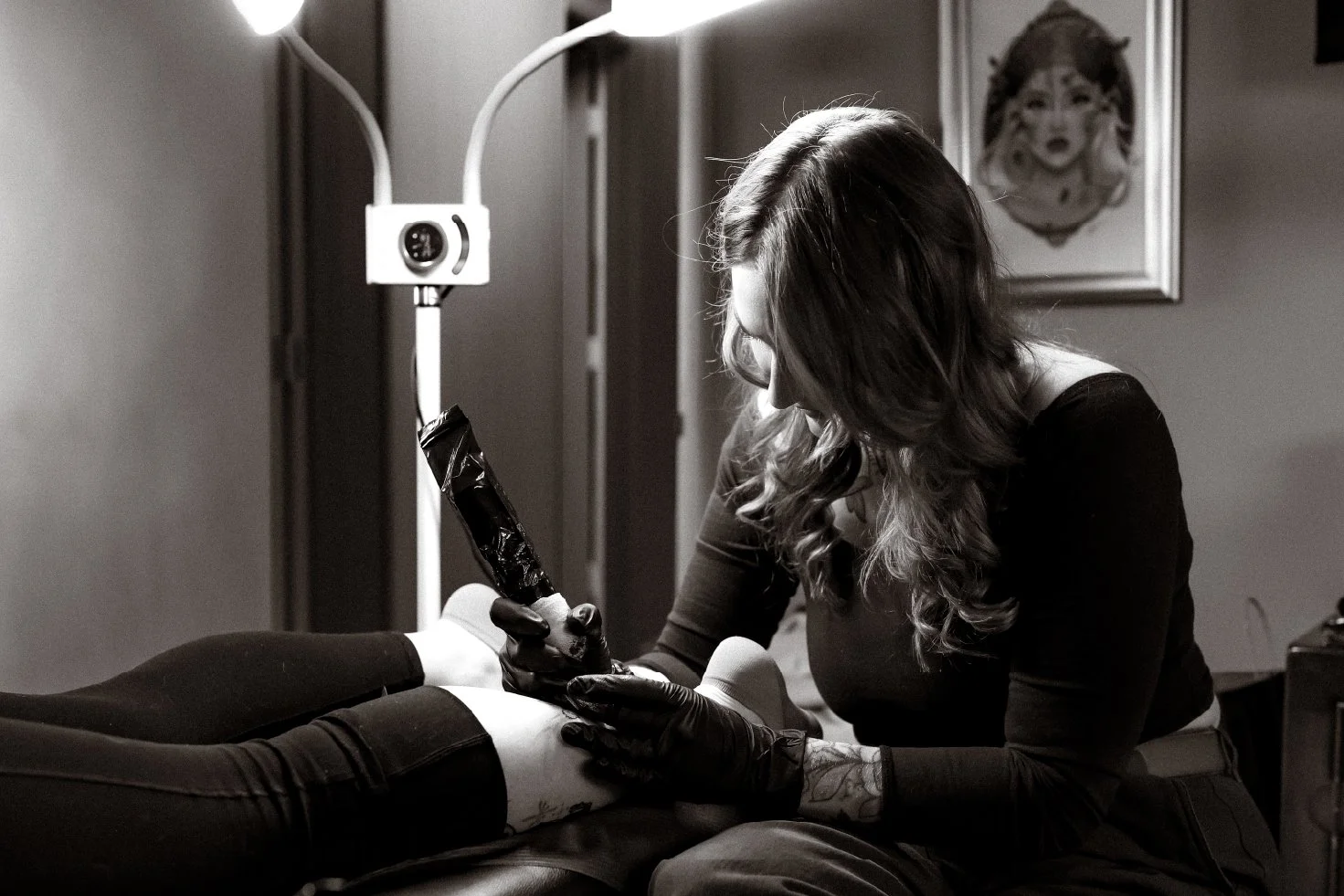 A woman with long hair, wearing black gloves and black clothing, is tattooing a person's leg in a tattoo studio. The studio has framed artwork on the wall, and the woman is focused on her work.