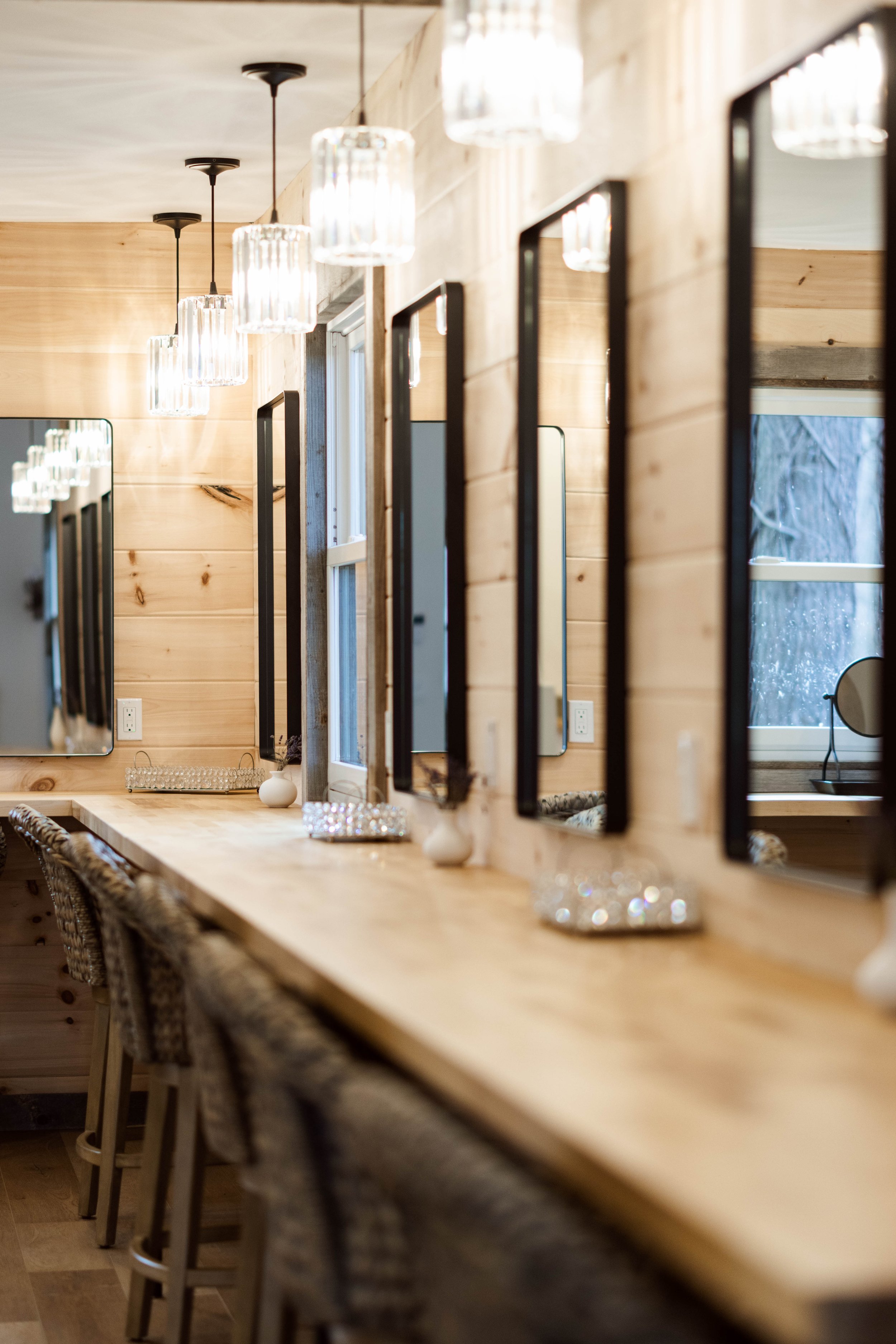 A wooden bathroom vanity with multiple mirrors, wicker chairs, and modern hanging light fixtures. A window showing a snowy outdoor scene is reflected in one of the mirrors.