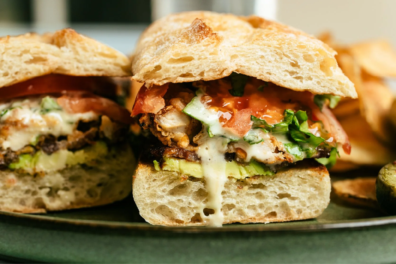 A close-up of a sandwich with melted cheese, grilled chicken, lettuce, tomato, and bacon inside a toasted bread bun, with a side of potato chips in the background.