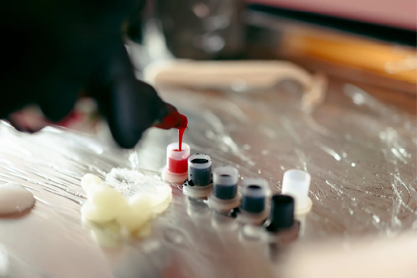 A person wearing black gloves filling a small bottle with red ink or dye in a lab setting, with multiple bottles of ink or dye lined up on a metallic surface.