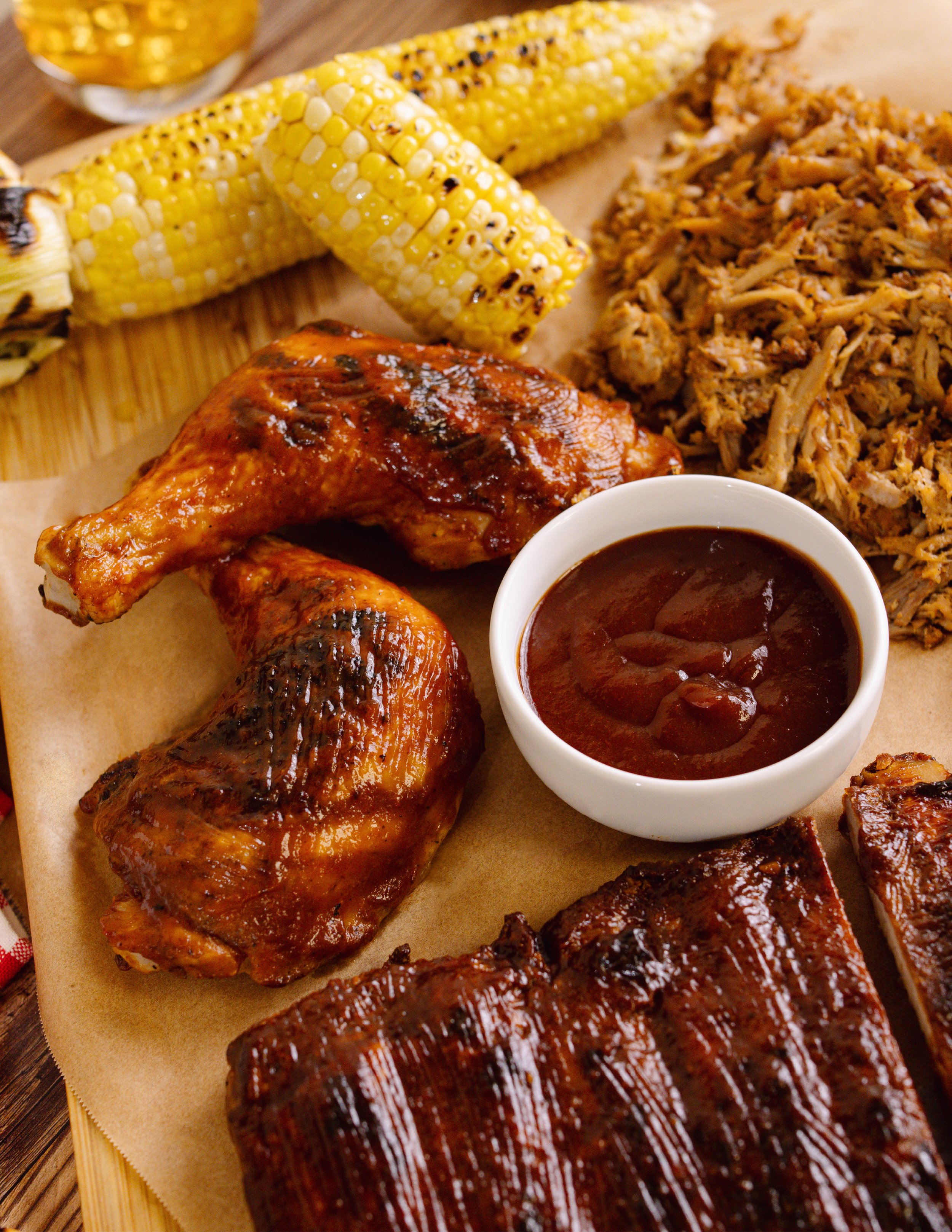 Close-up of smoked ribs, grilled corn on the cob, and a bowl of barbecue sauce on a wooden serving tray.
