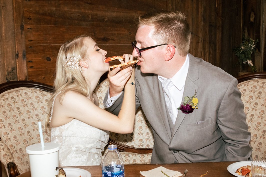 A young woman and a man in a gray suit sharing a slice of pizza at a wooden table.