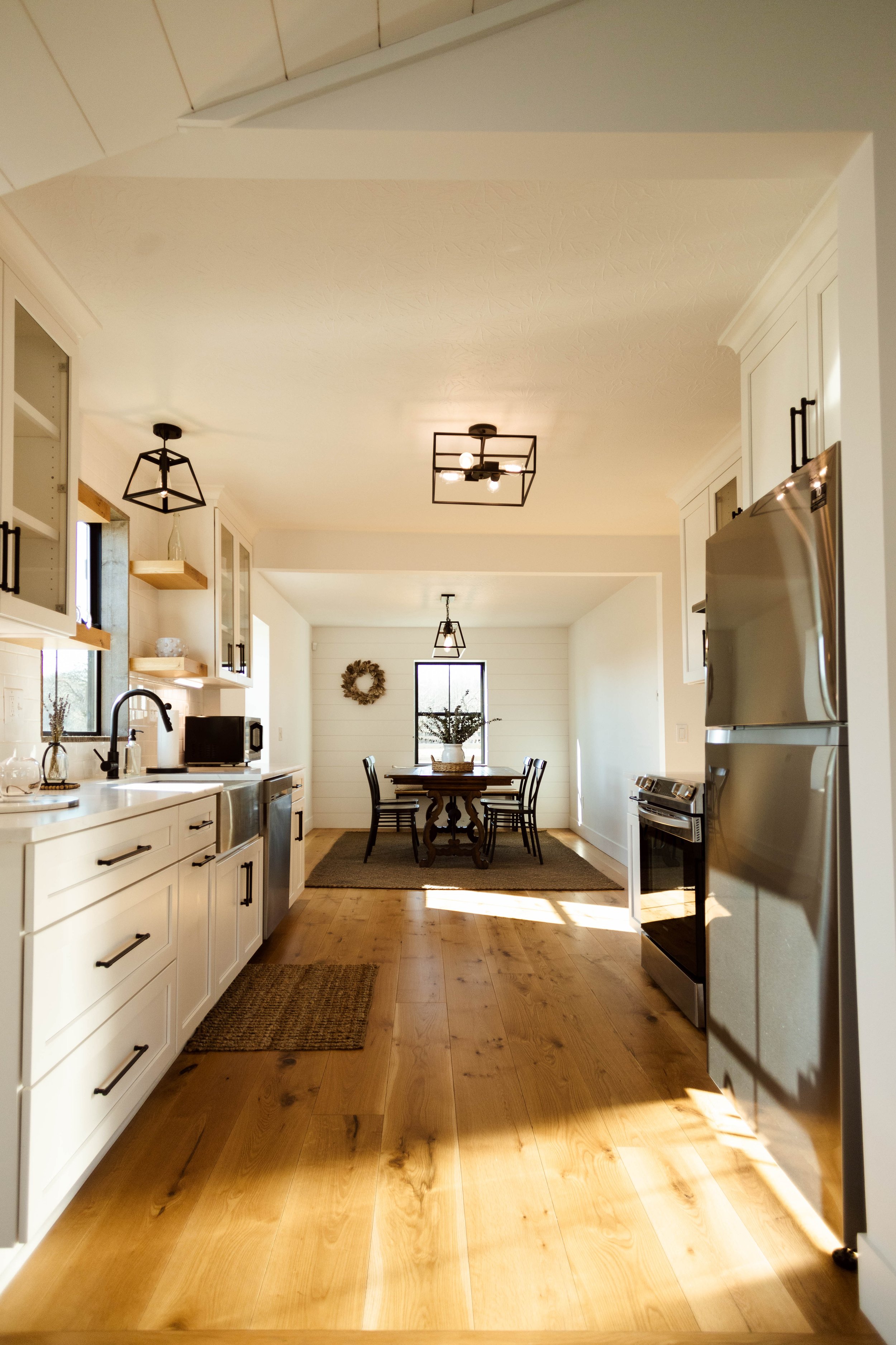 Bright, modern kitchen with white cabinetry, black hardware, stainless steel appliances, wood floors, and a dining area with a dark wood table and six chairs, decorated with a wreath on the wall and a centerpiece on the table.