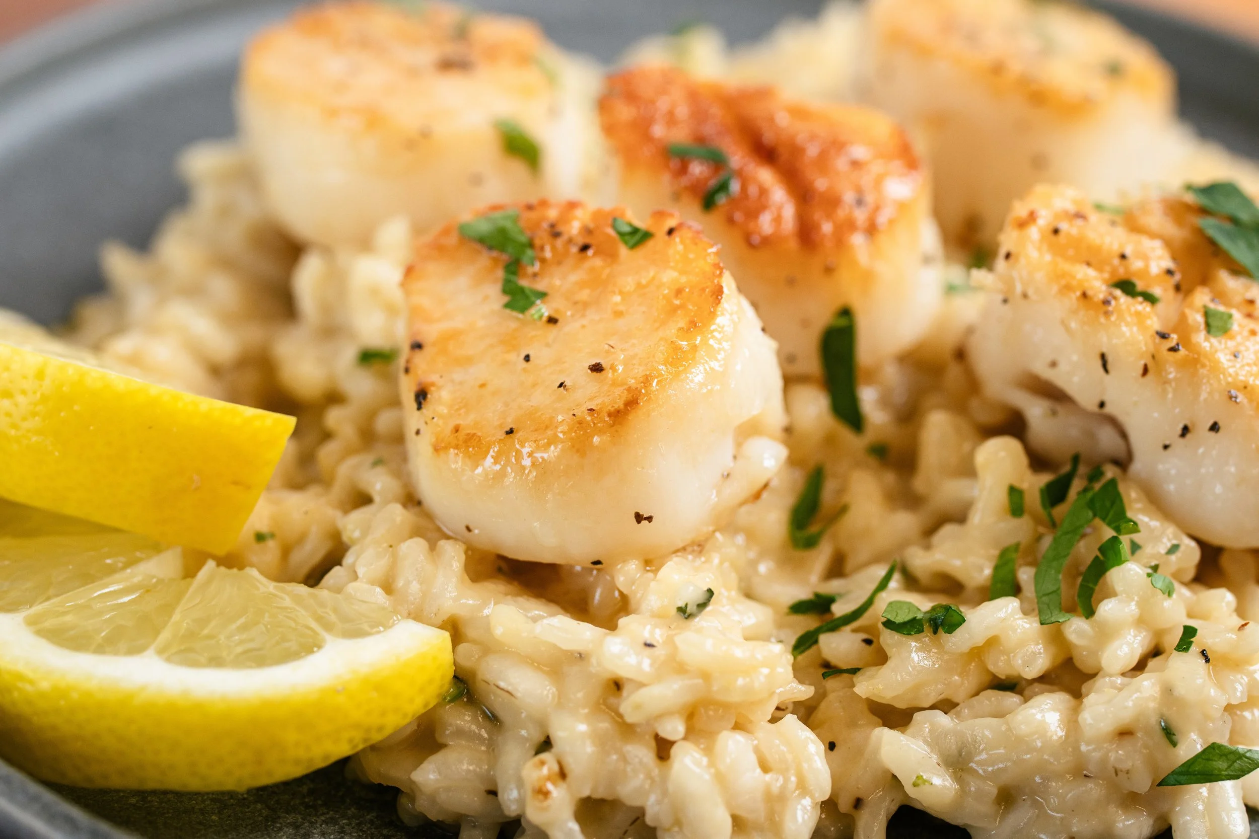 Close-up of seared scallops garnished with chopped herbs, served on a bed of creamy risotto, accompanied by lemon wedges on the side.