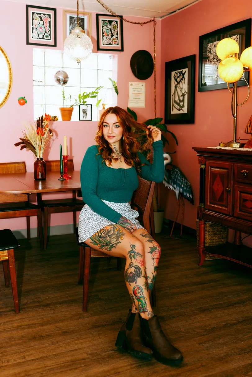 A young woman with red hair and tattoos on her legs, sitting on a chair in a pink-colored room decorated with artwork, plants, and lamps, while holding her hair.
