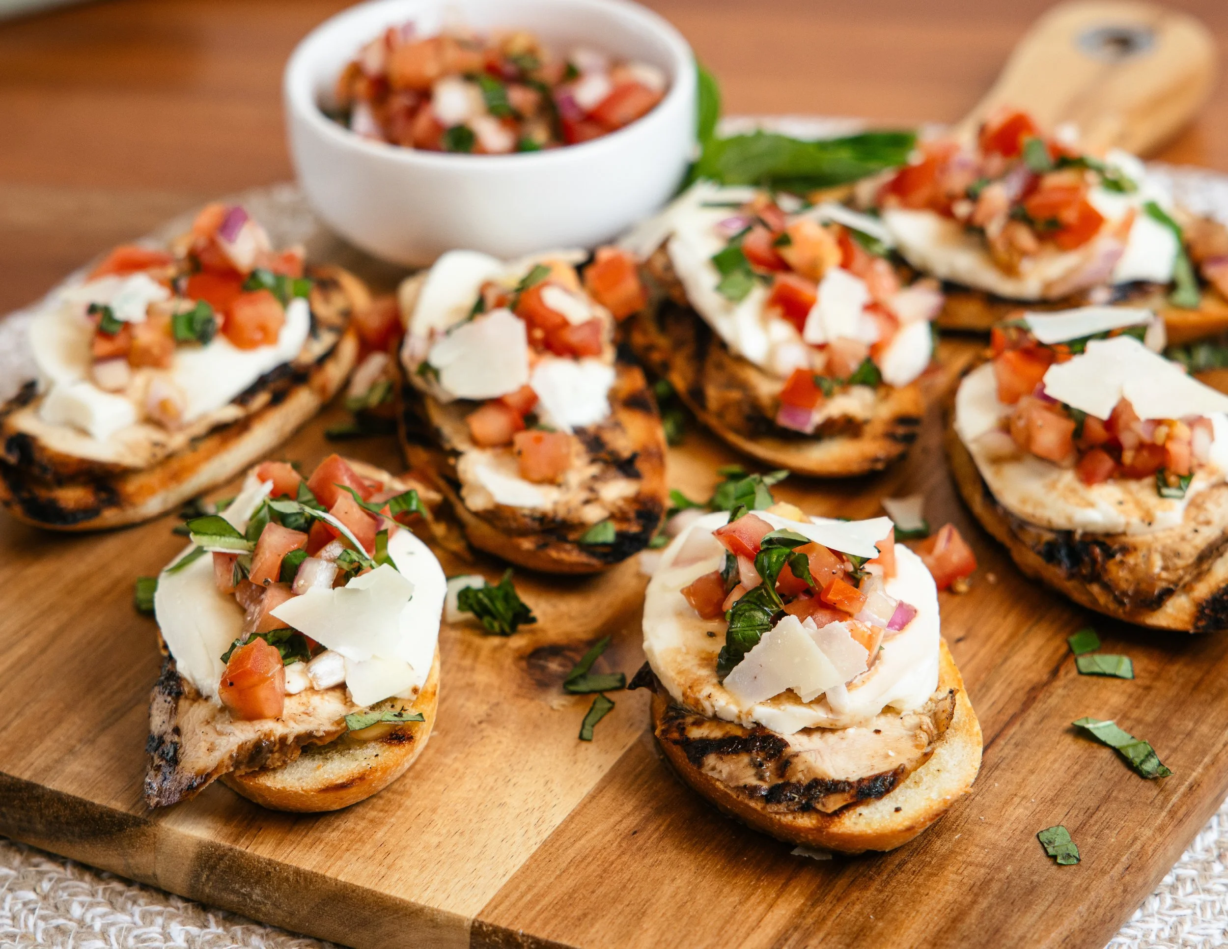 Grilled bruschetta topped with chopped tomatoes, onions, basil, and shaved cheese, served on a wooden board.