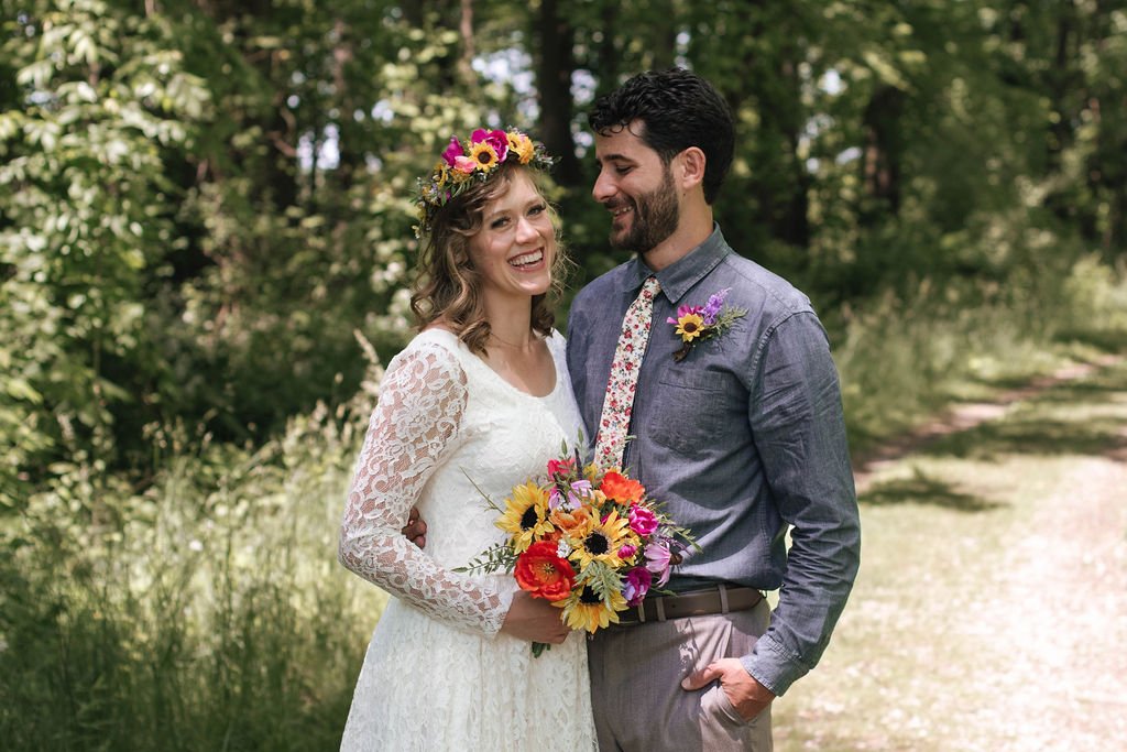 A bride and groom standing together outdoors, smiling, with the bride holding a colorful bouquet and wearing a floral crown, surrounded by green trees and foliage.