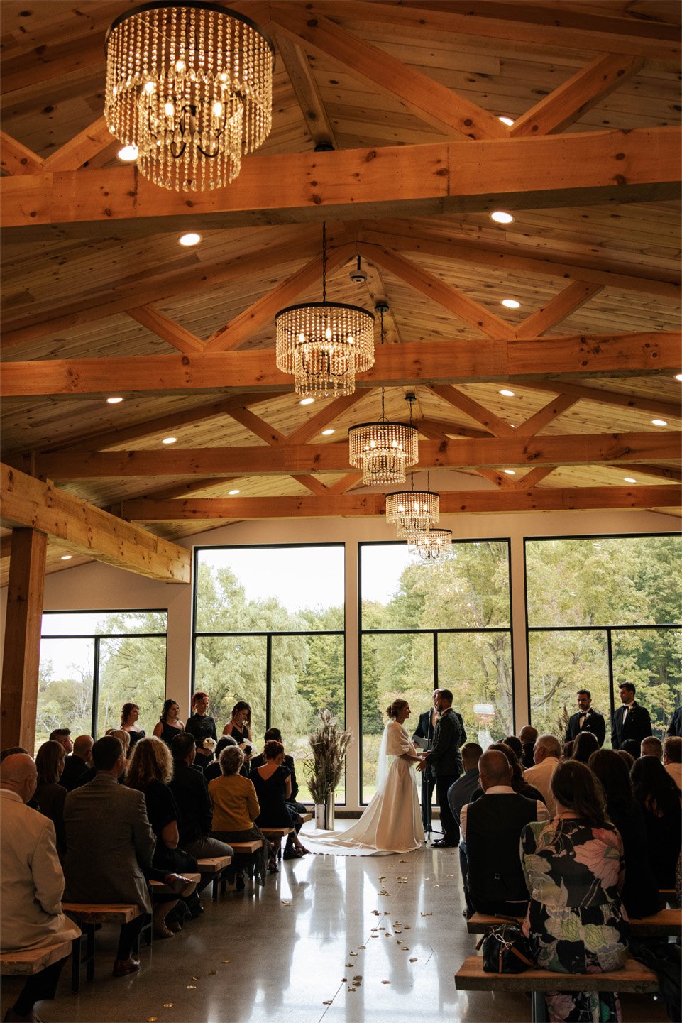 Indoor wedding ceremony with a bride and groom exchanging vows in front of seated guests, large windows showing trees outside, wood ceiling with chandeliers.