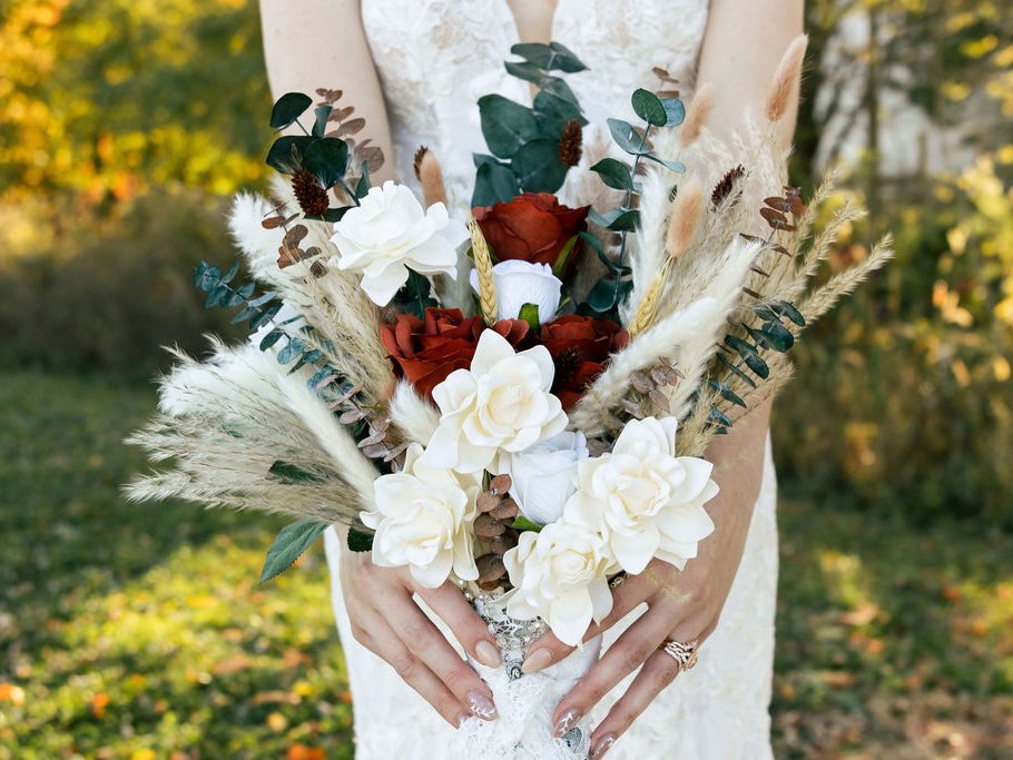 A woman in a white dress holding a bouquet of flowers with white, red, and beige blooms, surrounded by greenery and tall grasses, outdoors with a blurred background of trees and foliage.