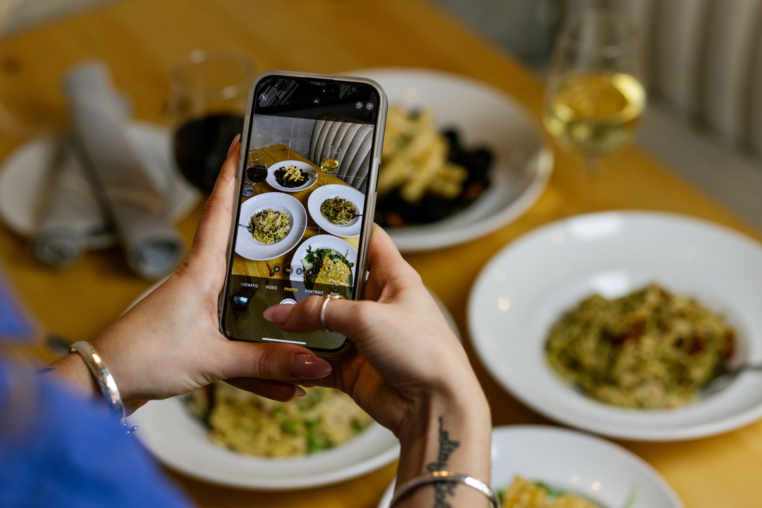 Person taking a photo of plates of pasta and wine on a wooden table.