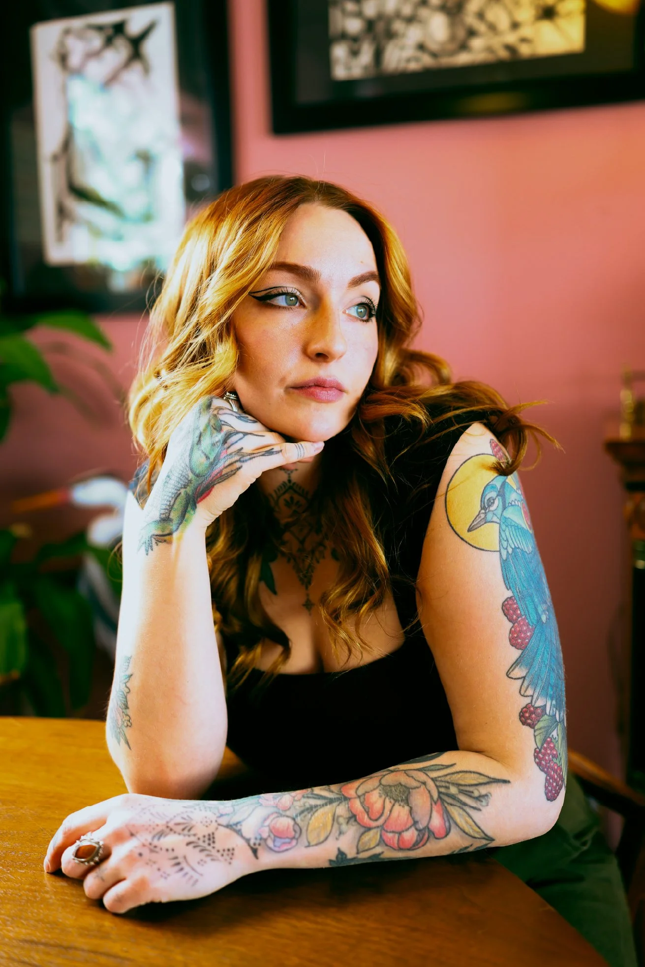 A young woman with long wavy red hair and blue eyes, wearing black clothing, is sitting at a wooden table with her left arm resting on it. She has extensive colorful tattoos on her arms, including a peacock, flowers, and other designs. She is looking