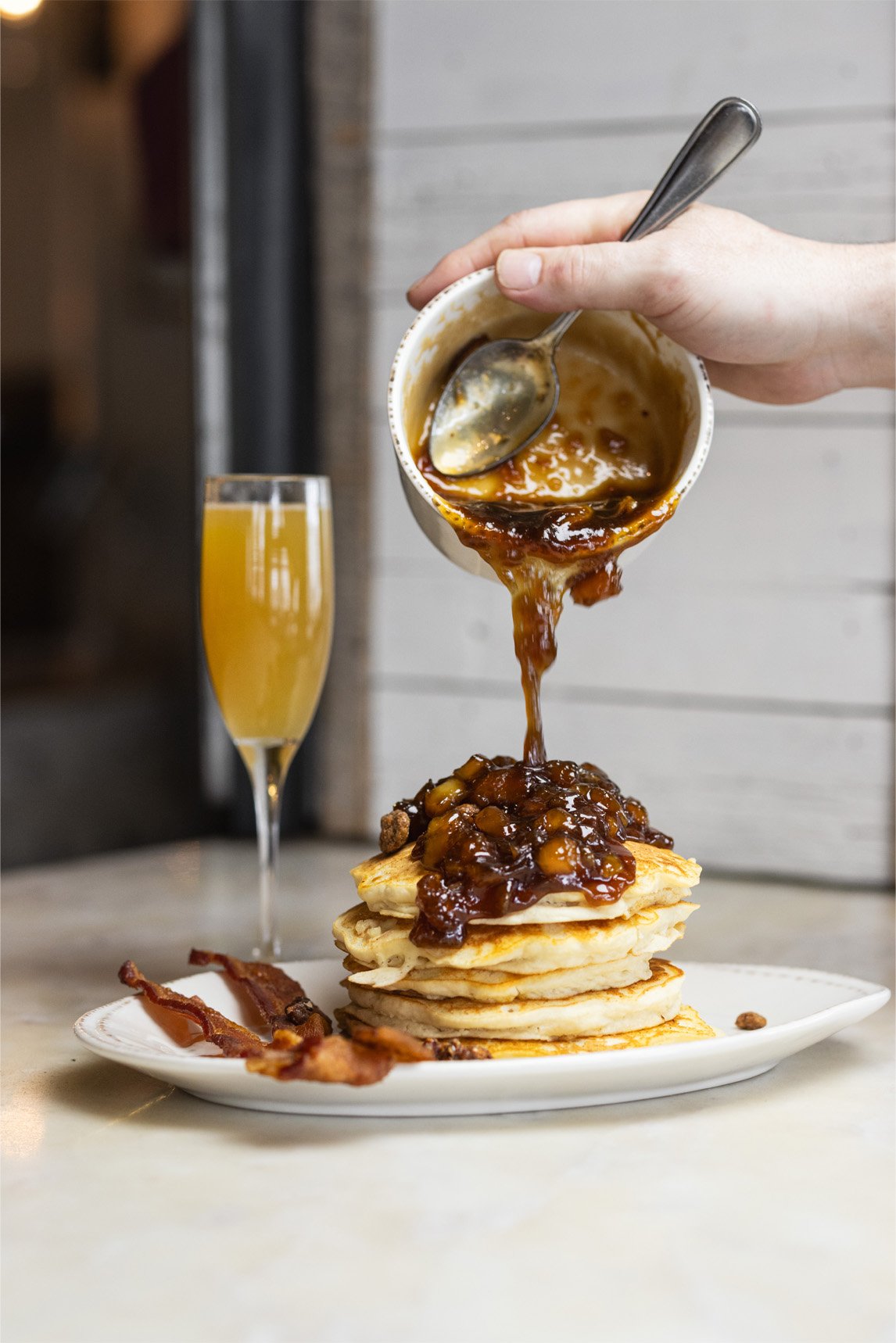 Stack of five golden pancakes topped with fruit-filled syrup, with bacon strips on the side and a glass of orange-colored beverage in the background. A hand pours gravy or sauce from a small bowl onto the pancakes.
