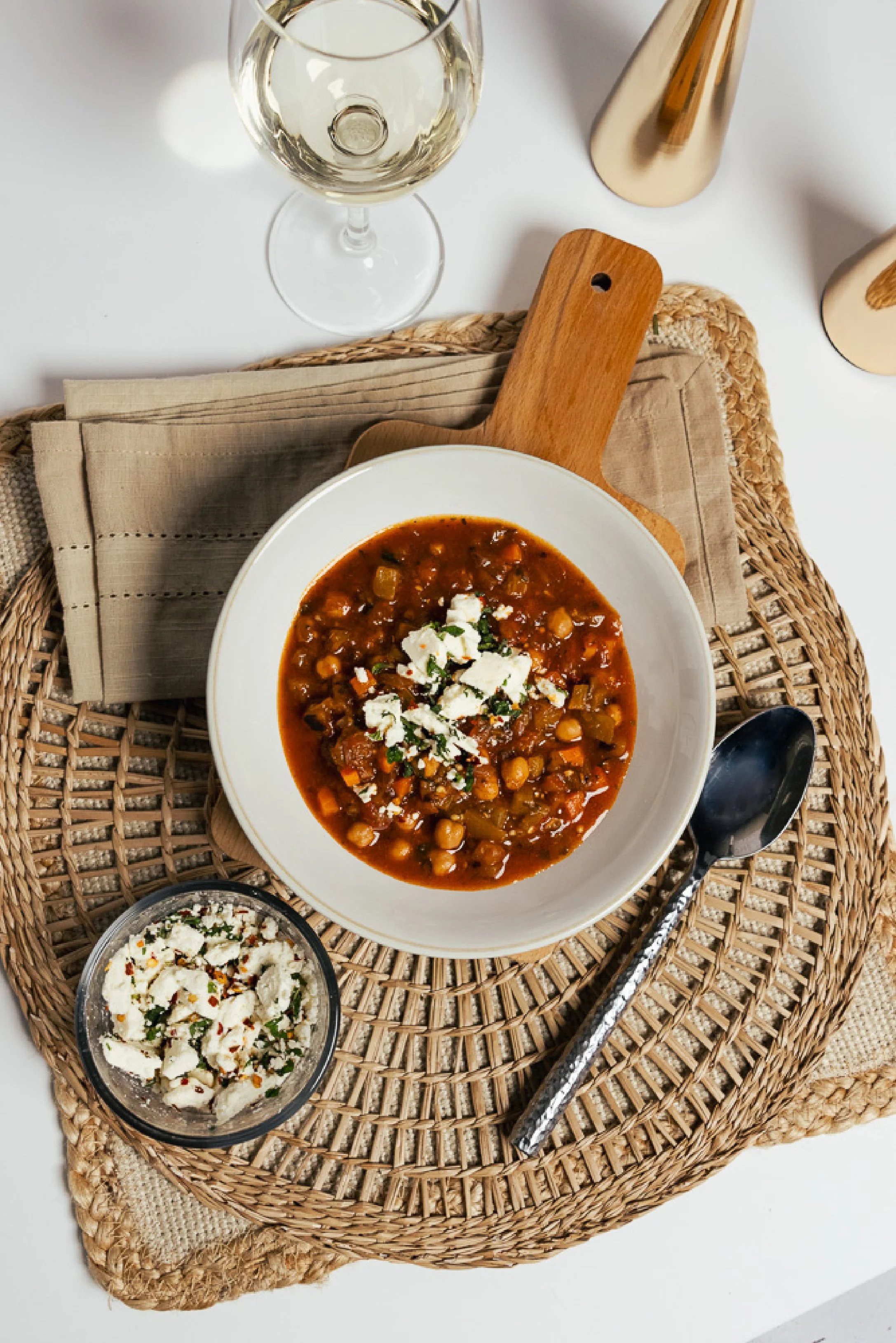 A bowl of chickpea stew garnished with crumbled cheese and herbs on a wicker placemat, with a glass of white wine and a small bowl of salad nearby.