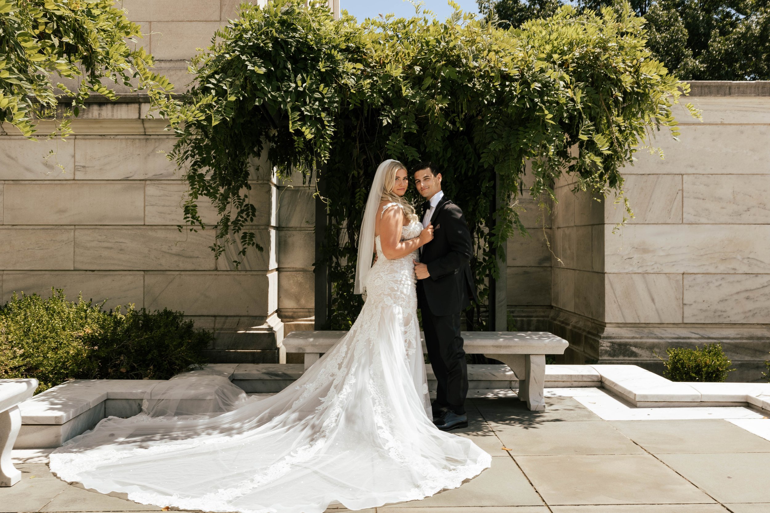 A bride in a white wedding gown with a long train and veil, and a groom in a black tuxedo, standing close together on a paved outdoor area with greenery and stone benches, under an arbor with leafy vines.