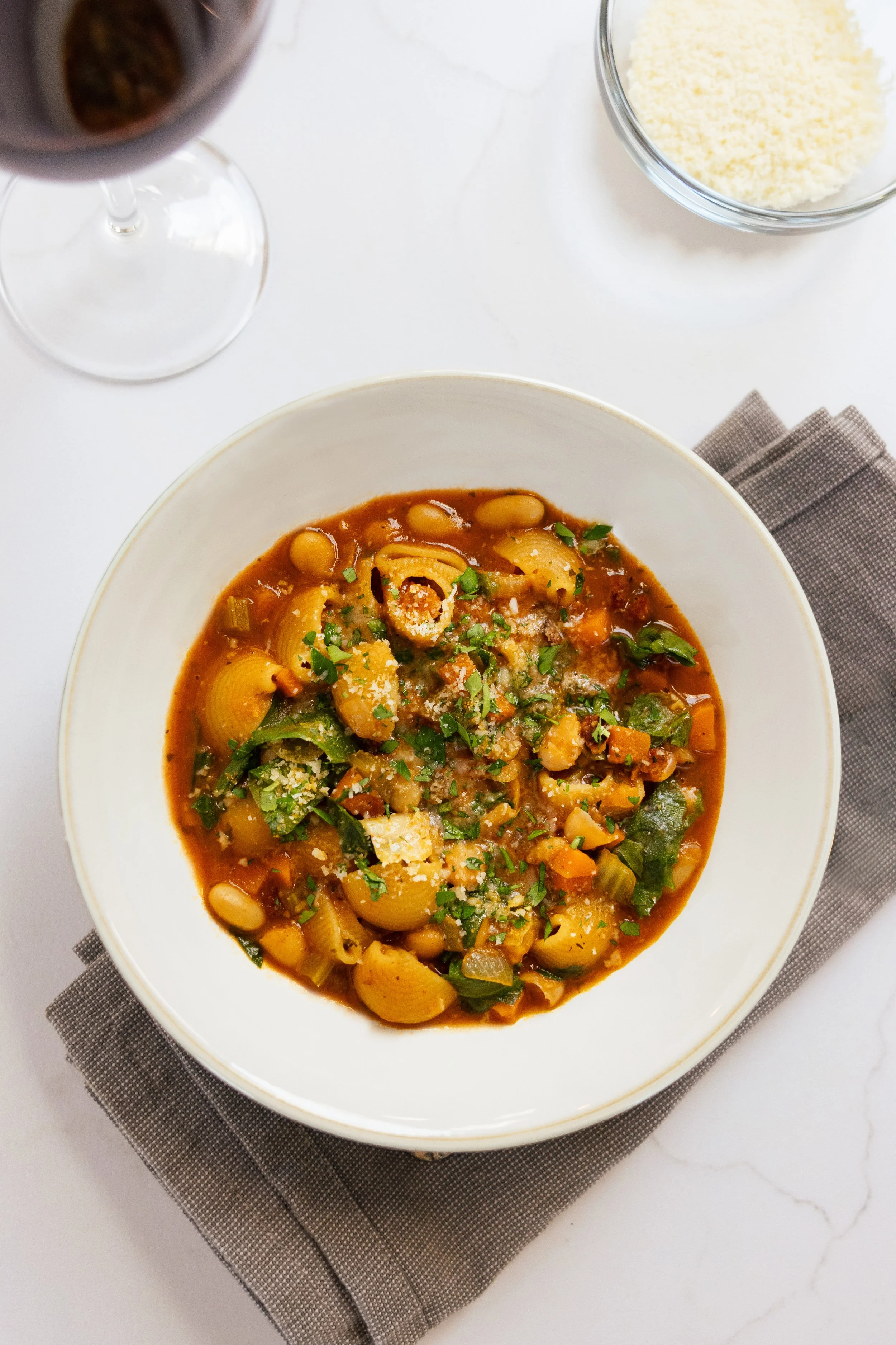 A bowl of pasta and beans stew garnished with chopped parsley and grated cheese, with a glass of red wine and a bowl of grated cheese on the white table.