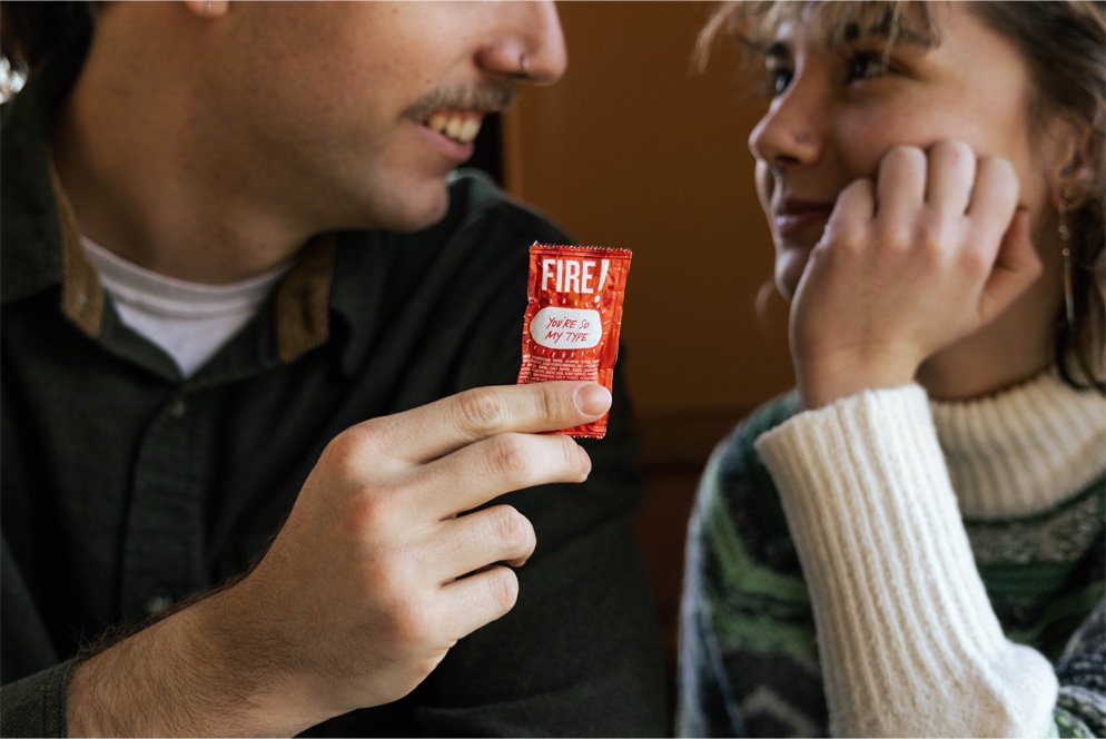 A man offering a red fire safety device labeled 'FIRE!' to a woman, who is smiling and resting her chin on her hand.
