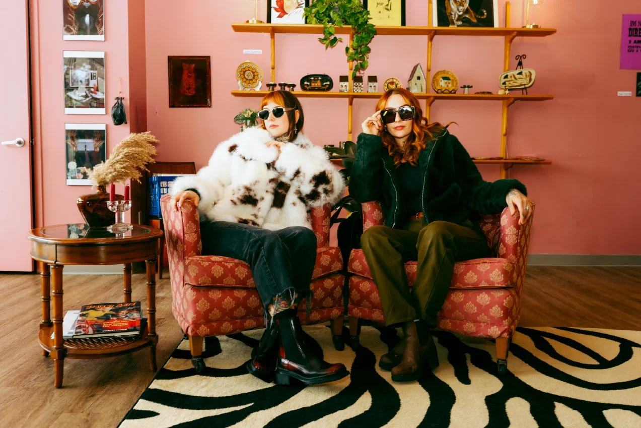 Two women sitting on pink patterned armchairs in a room with pink walls, one wearing a white fur coat and sunglasses, and the other wearing a black coat and sunglasses, with a black and white zebra print rug on the floor, and shelves with decorative 