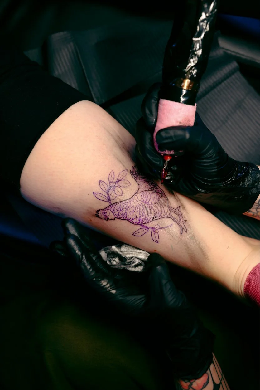A person with a tattoo on their thigh of a chameleon surrounded by leaves, as they are getting the tattoo inked by a tattoo artist wearing black gloves.
