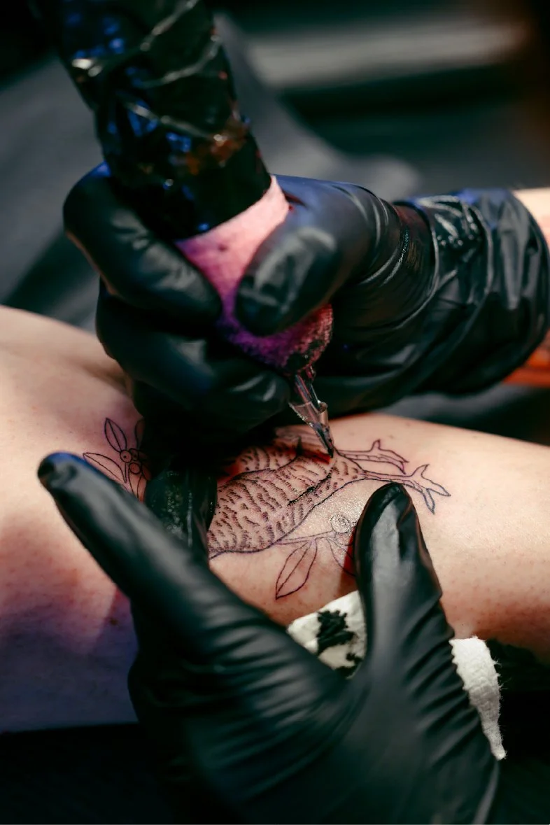 A tattoo artist in black gloves is creating a detailed floral tattoo on a person's thigh. The artist is using a tattoo machine, and the tattoo features a large flower surrounded by smaller floral elements.