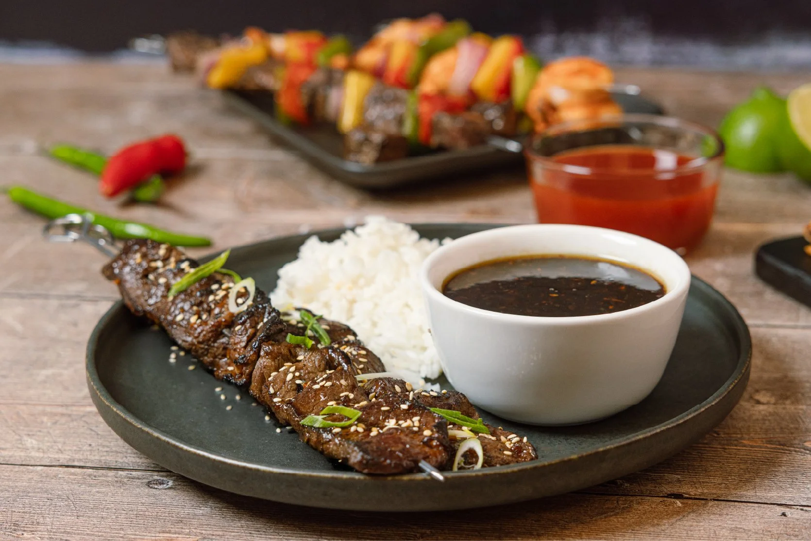 Plate with grilled beef skewer topped with sesame seeds and green onions, served with white rice and dark dipping sauce, on a black plate. In the background, there are various grilled skewers, a small bowl of red sauce, a lime, and green chili pepper