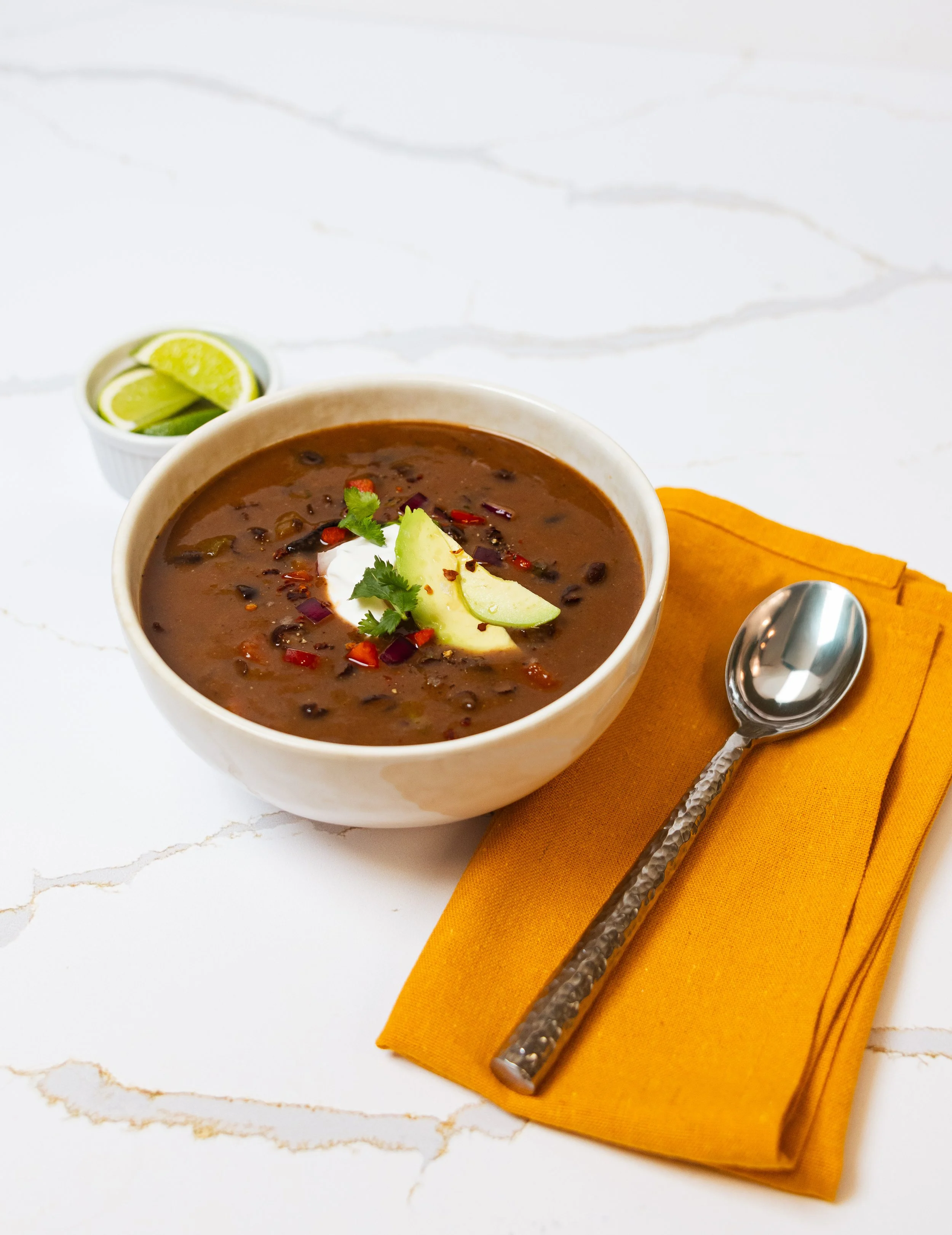 A bowl of spicy black bean soup garnished with avocado slices, cilantro, and red chili flakes, with lime wedges in a small white bowl, placed on an orange napkin with a silver spoon on a white marble surface.