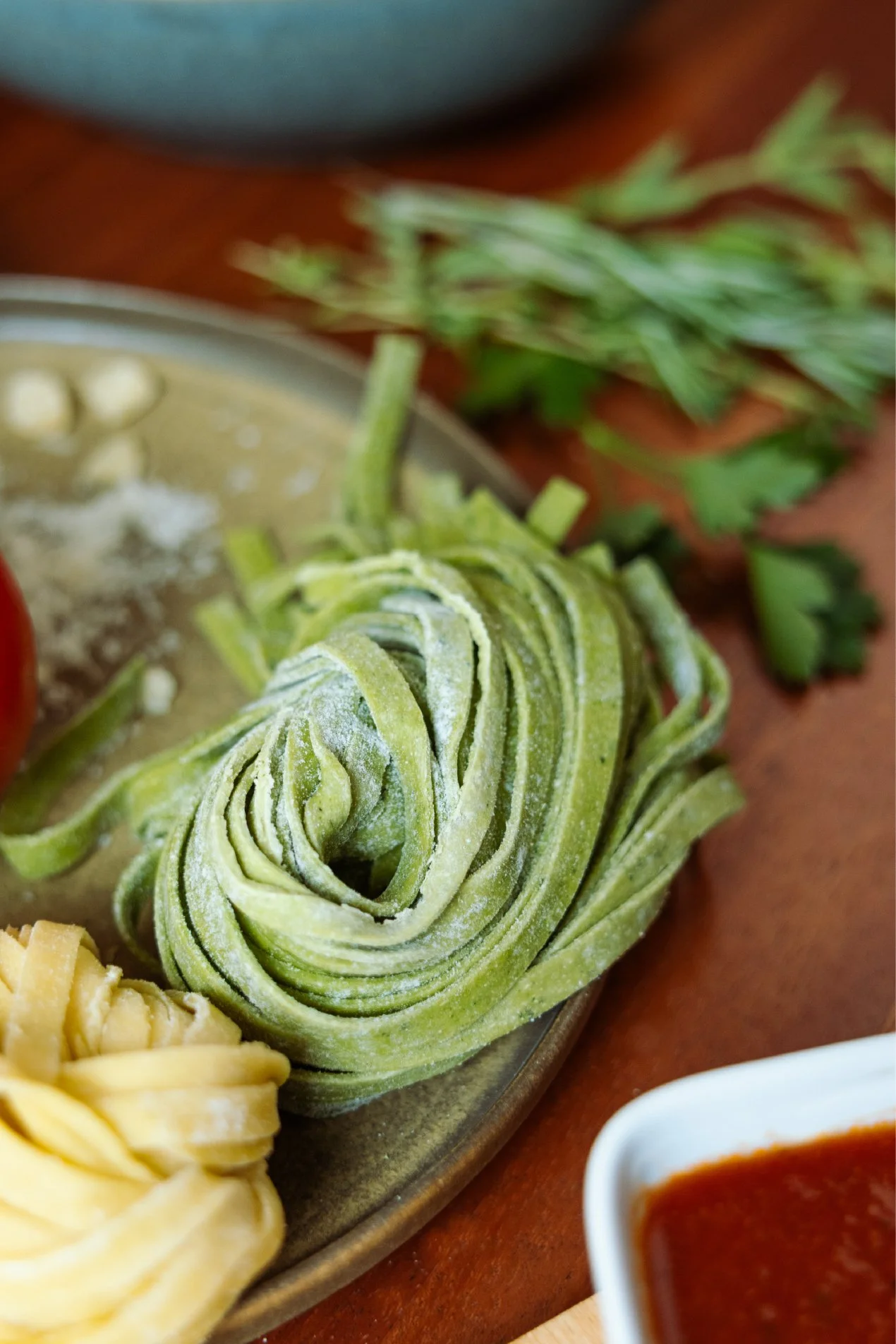 A close-up of uncooked green spinach pasta fettuccine on a wooden board accompanied by a small dish of red marinara sauce.