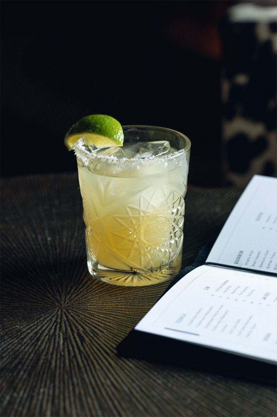 A glass of margarita with a lime wedge and salted rim on a dark textured table next to an open menu.
