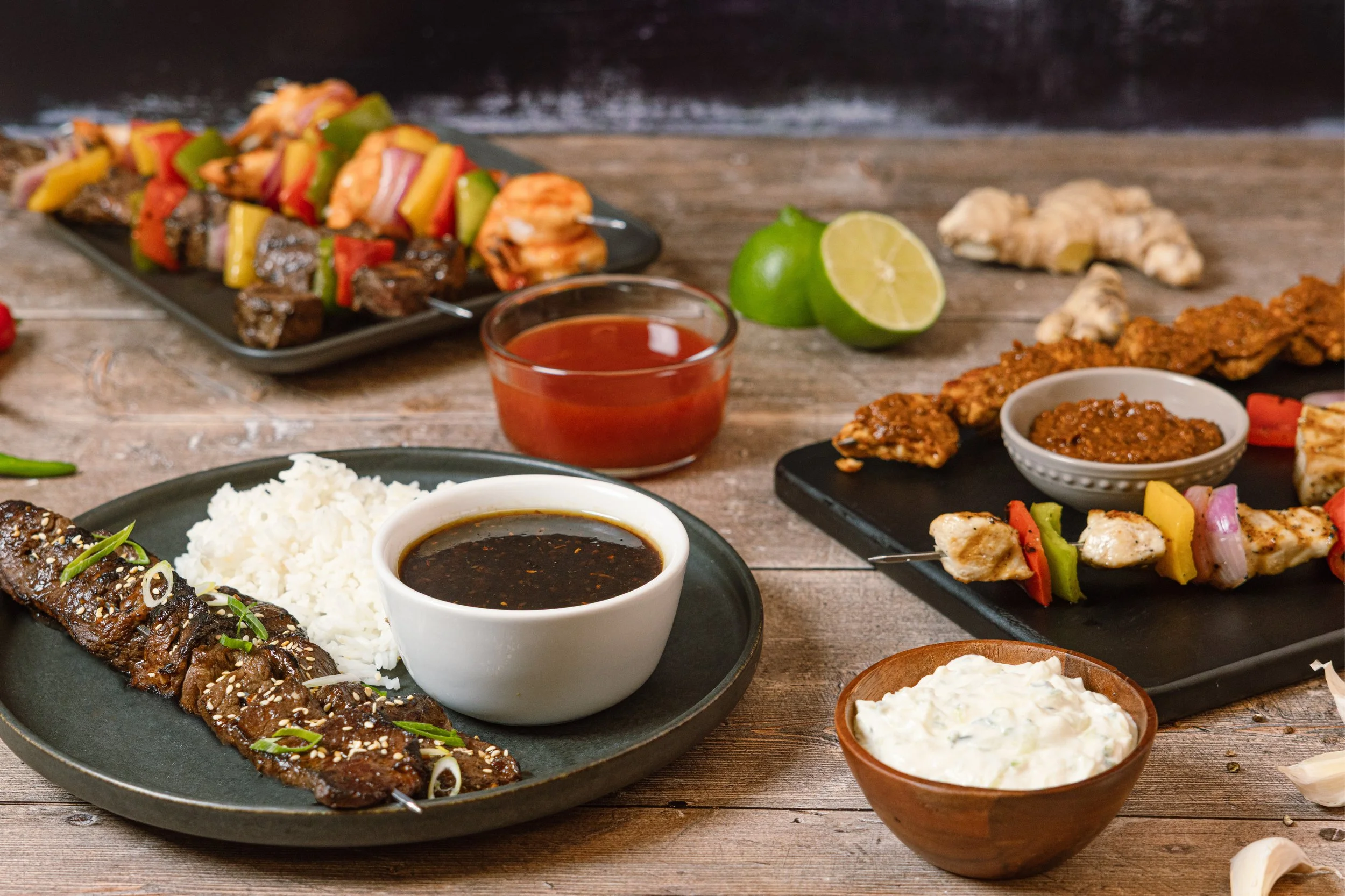 A variety of Asian cuisine dishes on a rustic wooden table, including grilled meat skewers, rice, dipping sauces, fresh lime, ginger, and vegetable skewers.