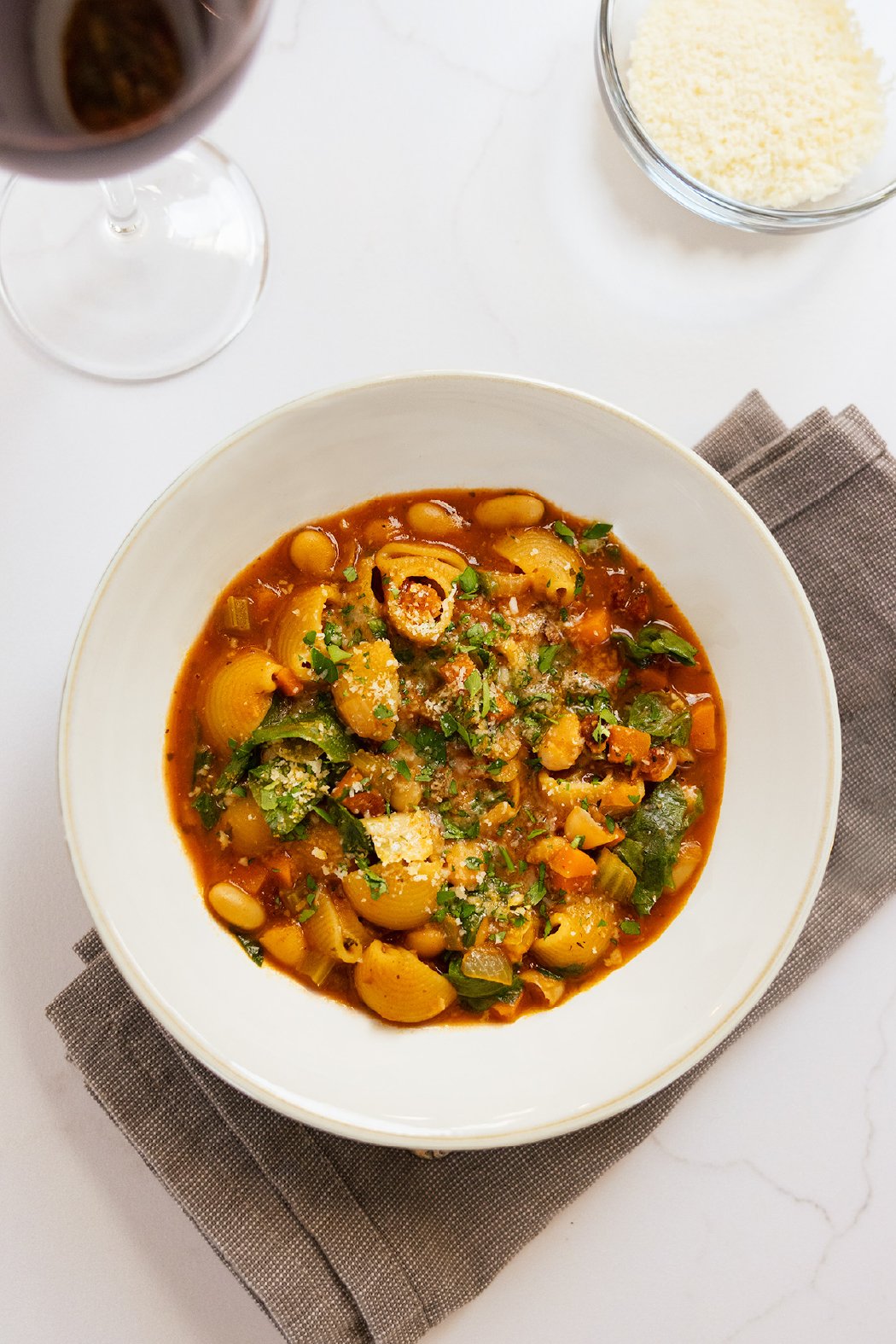 A bowl of vegetable minestrone soup with pasta, beans, carrots, celery, and greens, garnished with parmesan cheese and chopped herbs, on a gray napkin next to a glass of red wine and a bowl of grated cheese.