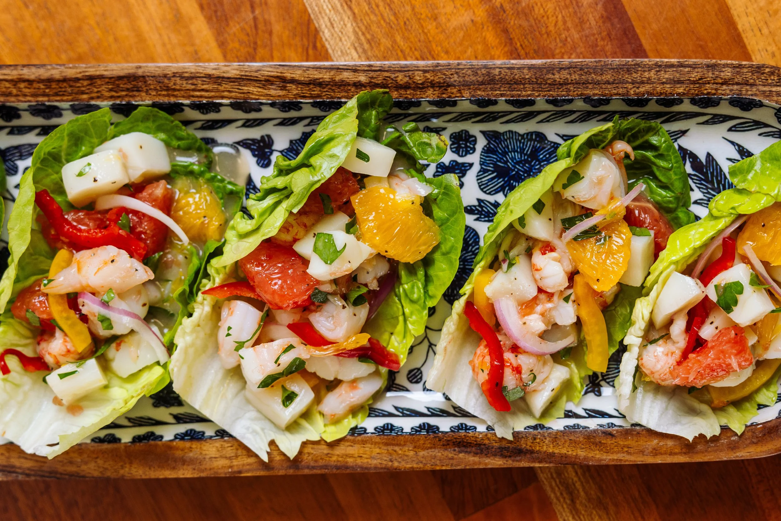 Close-up of four lettuce cups filled with chopped seafood, diced vegetables, and herbs, on a decorative rectangular tray with black and white floral patterns, on a wooden table.