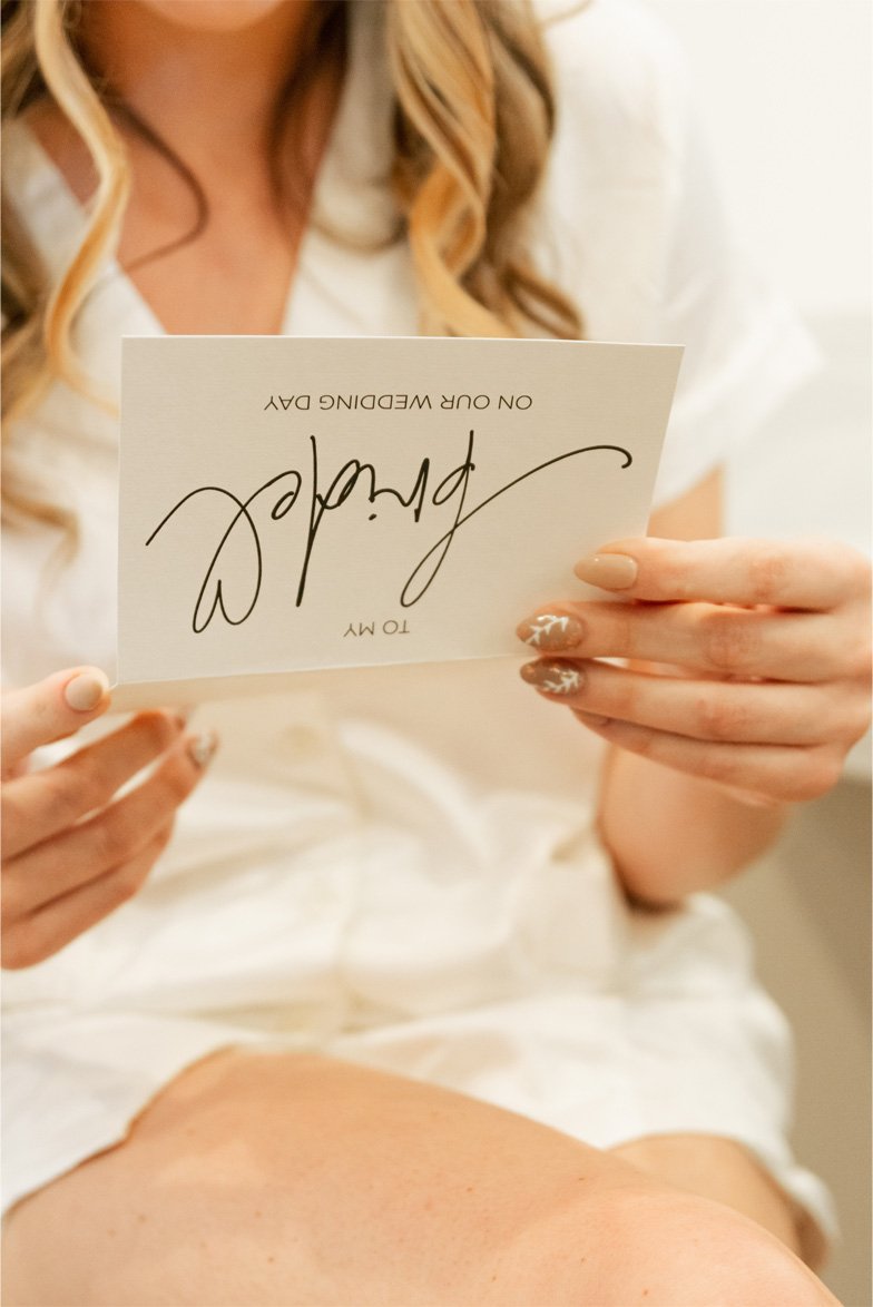 A woman in white clothing holding a wedding card with text 'Tom, on our wedding day,' with her nails painted in a beige and white floral pattern.