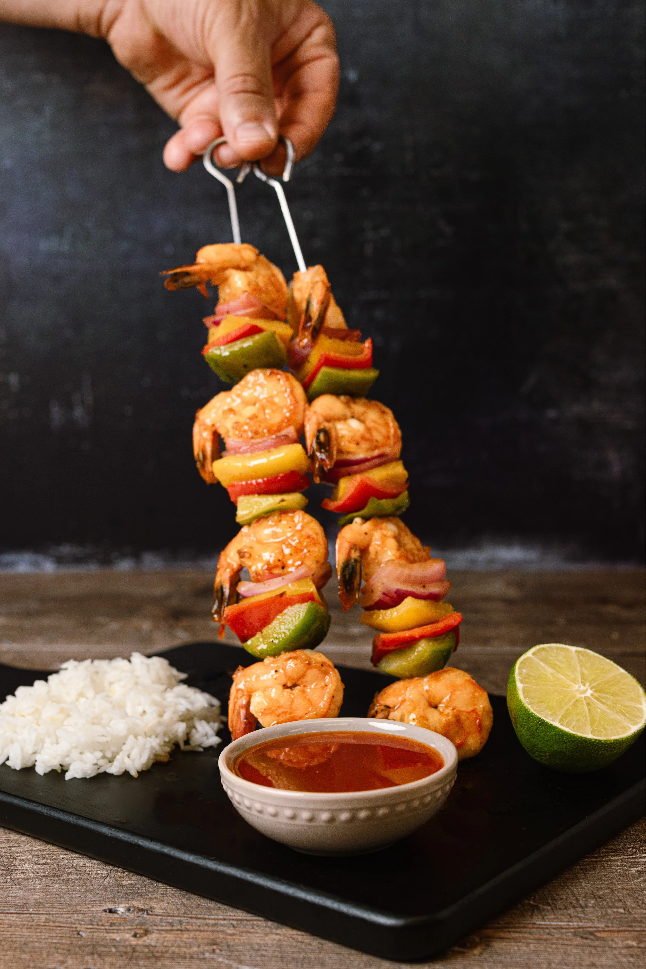 Close-up of skewered shrimp and colorful vegetables on a black platter, with white rice, a lime half, and a small bowl of dipping sauce on a rustic wooden surface.