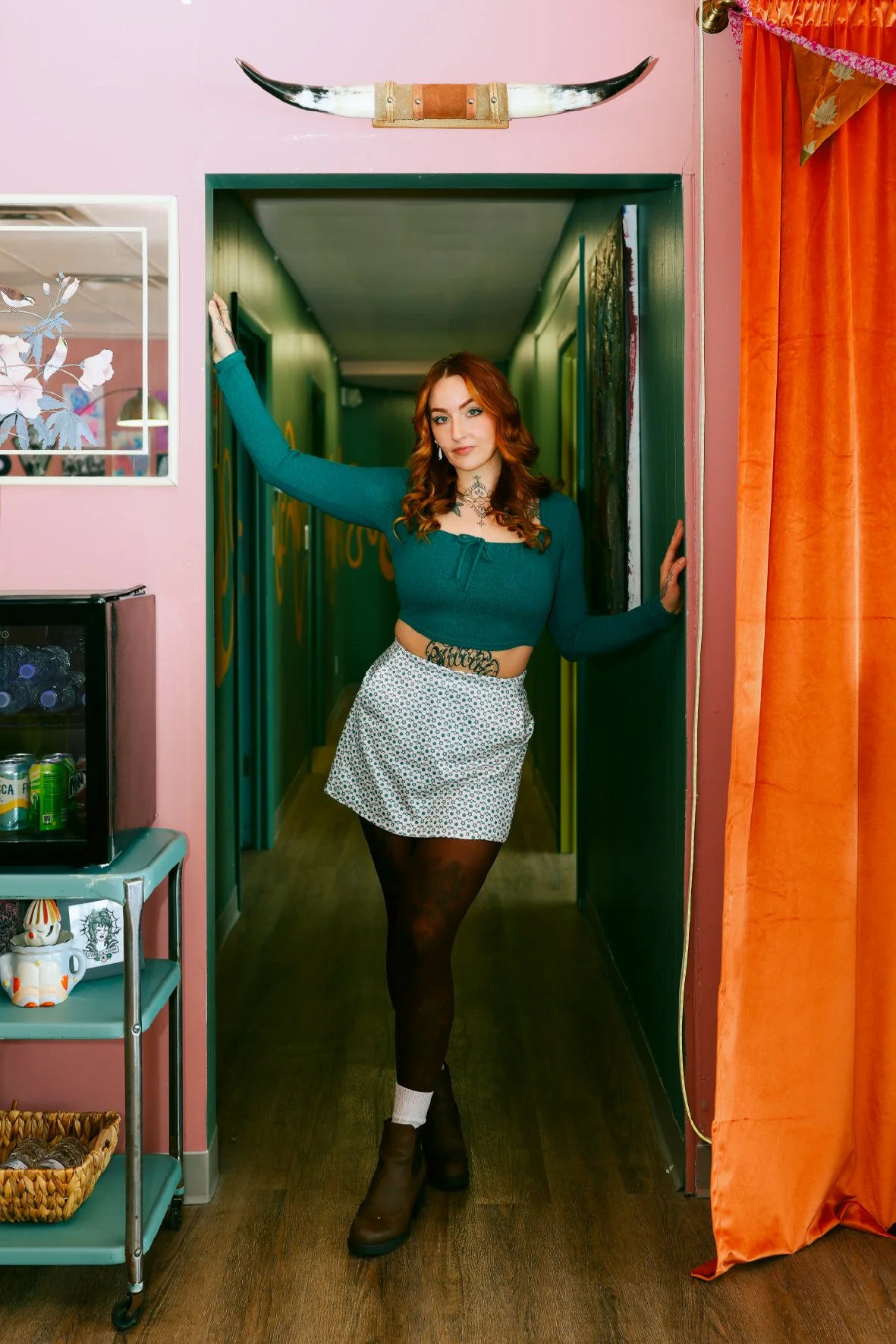 A woman with red hair and tattoos posing in a doorway in a colorful room. She is wearing a teal long-sleeve crop top, a white skirt with black polka dots, black tights, and brown boots.