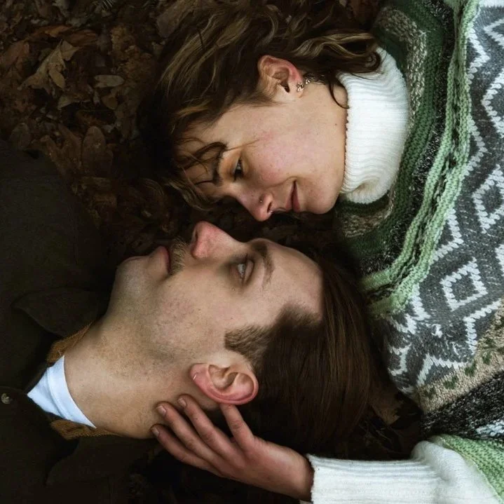 A young woman lying on the ground, gently touching foreheads with a young man, both with eyes closed, sharing an intimate moment surrounded by leaves.