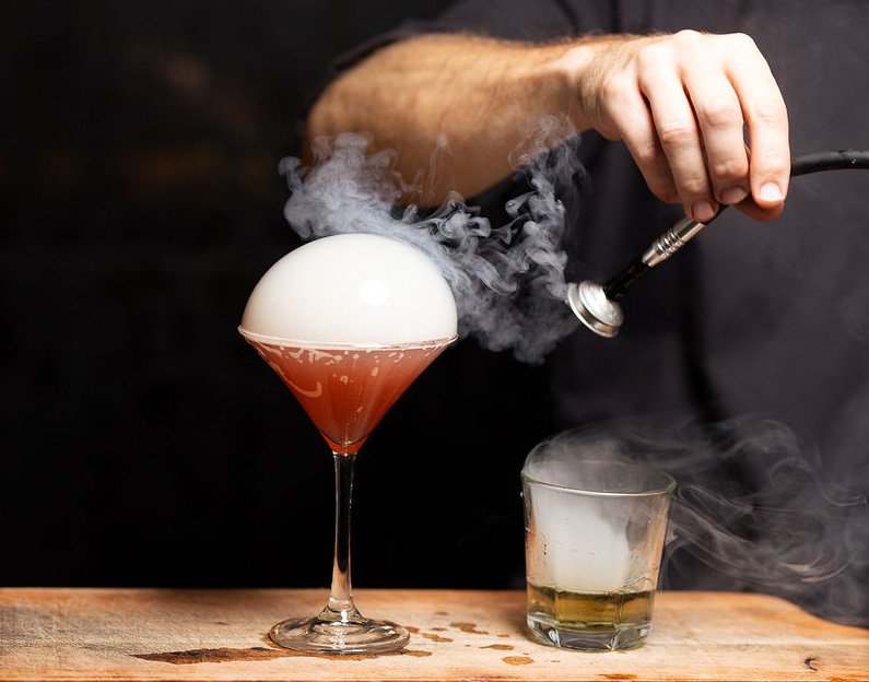 A bartender uses a smoking gun to create dry ice fog inside a cocktail glass with a frothy pink drink, with a smaller glass containing smoke beside it on a wooden surface.