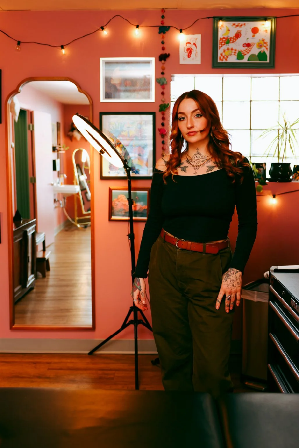 A woman with long auburn hair and tattoos on her neck and hands stands in a warmly-lit room decorated with colorful artwork, string lights, and a window. She is wearing a black top and olive green pants with a brown belt, and looks directly at the ca