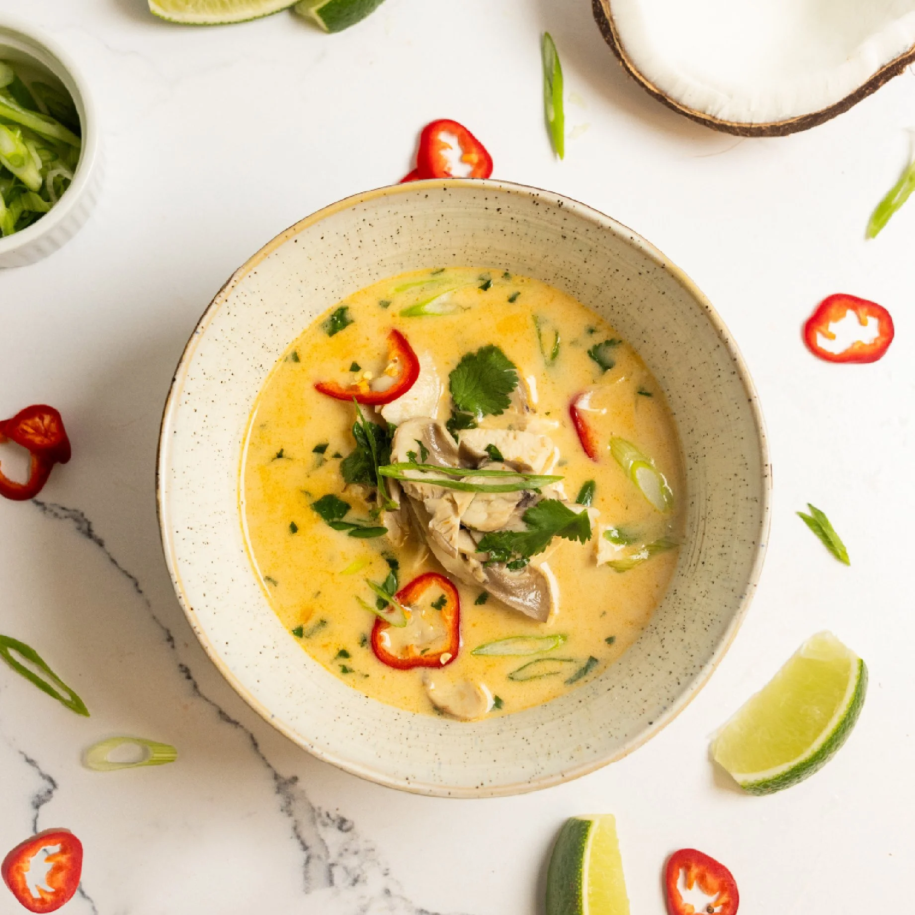 A bowl of Thai green curry with chicken, red chili slices, green onions, and cilantro, with lime wedges and coconut and chopped vegetables around it.