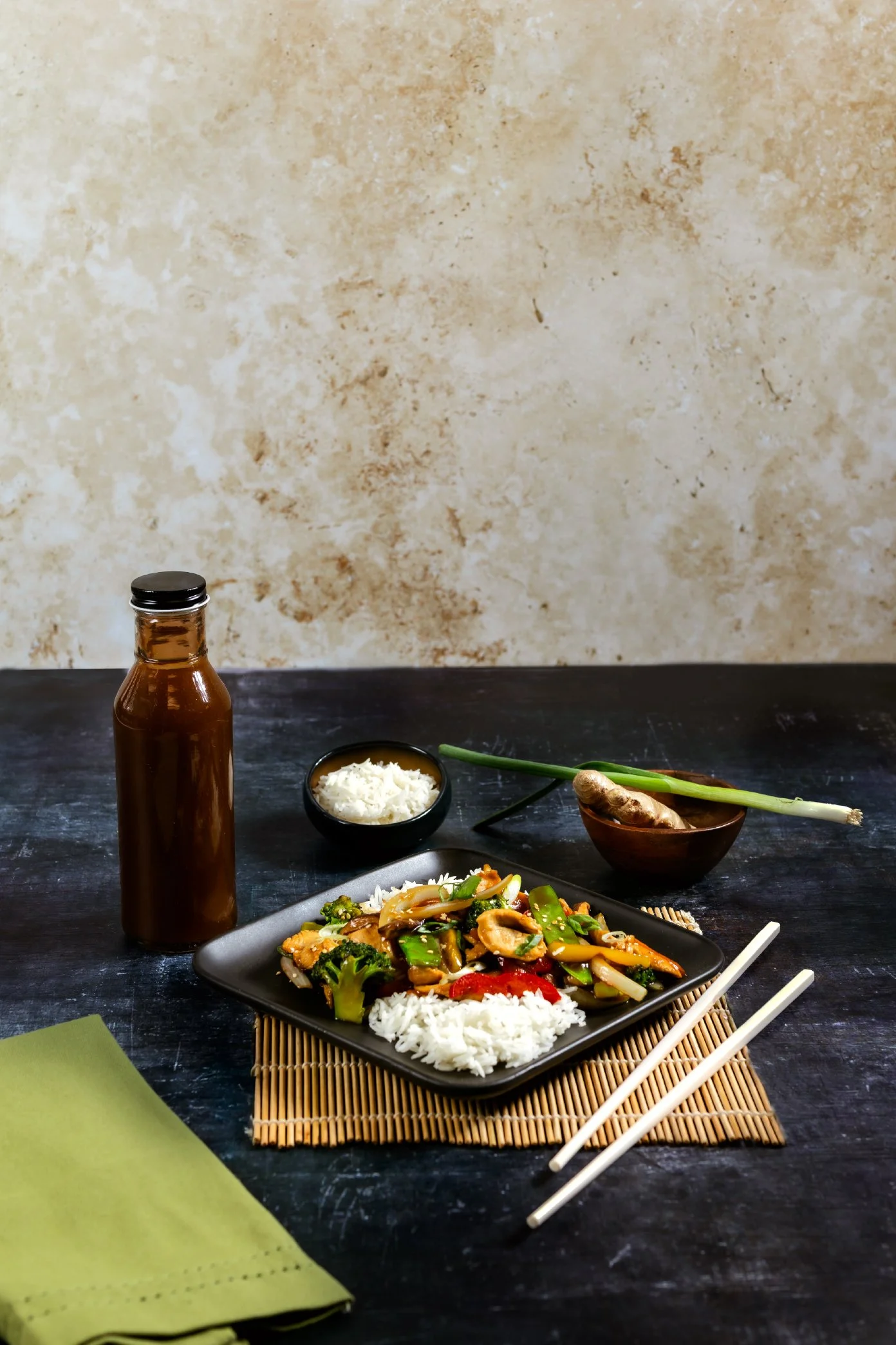 A black square plate with stir-fried vegetables and rice on a bamboo placemat, chopsticks, a glass soy sauce bottle, a small bowl of shredded white cheese, a small bowl with a ginger root, green onions, and a partially visible green napkin on a black