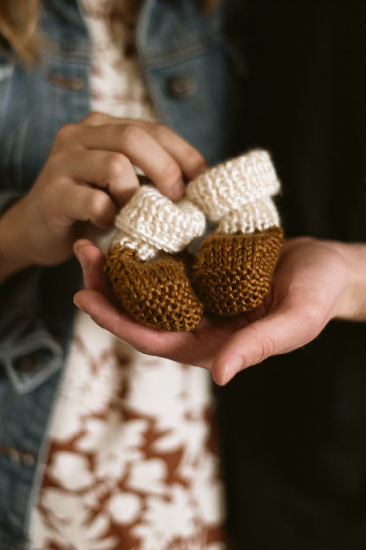 Person holding a pair of small knitted baby booties, one beige and one brown, outdoors.