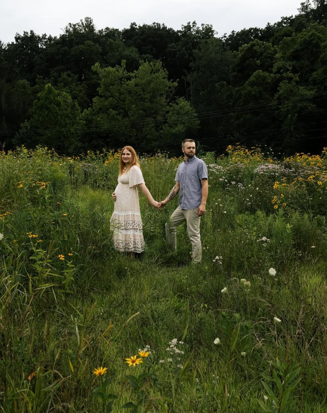 A man and a woman holding hands in a lush green field with flowers, with dense trees in the background.