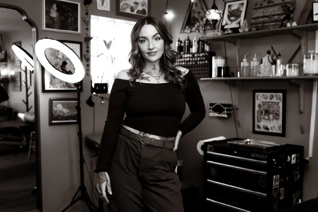 A woman with wavy hair and tattoos wearing a black off-shoulder top and dark pants, standing in a tattoo studio surrounded by artwork, tattoo supplies, and studio equipment.