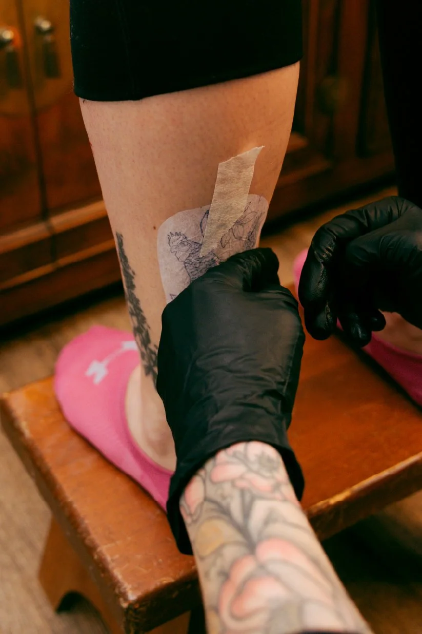 A person with tattoos on their arm is applying a tattoo transfer to their thigh, which is supported on a wooden stool. The person is wearing black gloves and pink socks.
