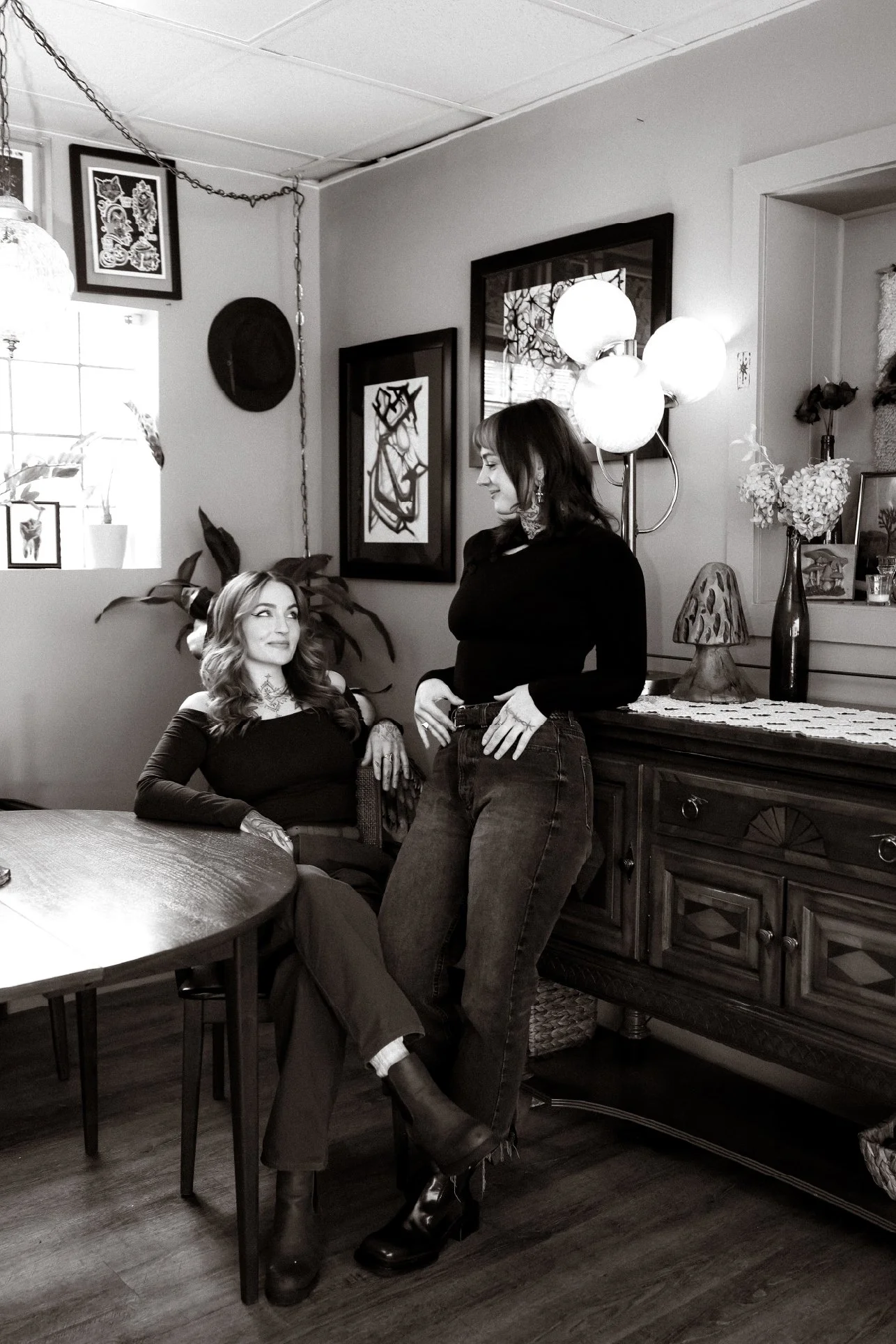 Two women in a cozy, decorated living room. One woman is seated at a wooden dining table, looking up at the other woman, standing near a sideboard with flowers. The standing woman has short hair and is smiling. The room has framed artwork on the wall