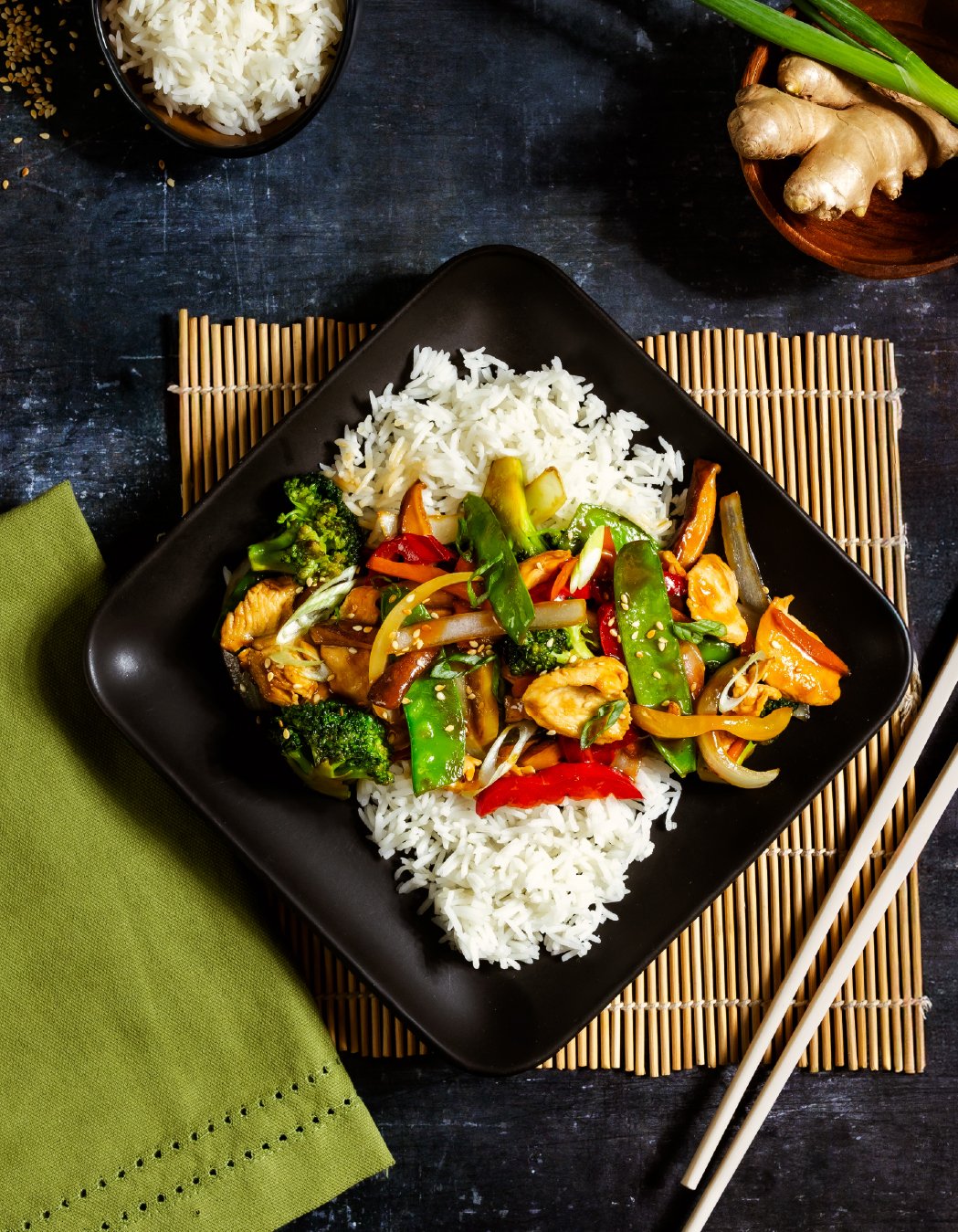 A black square plate with stir-fried vegetables and chicken served with white rice, chopsticks on the side, on a bamboo placemat. The vegetables include broccoli, snap peas, bell peppers, and onions. In the background, there is a bowl of rice, ginger
