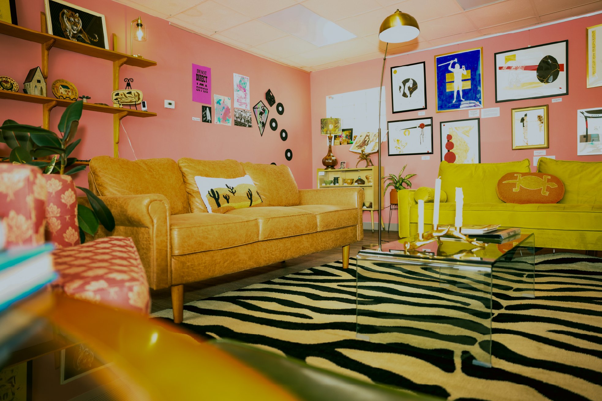 A living room with pink walls, decorated with modern art and wall decor. It features a mustard yellow sofa with a cactus pillow, a lime green cushion, and a black and white zebra-patterned rug. There is a black glass coffee table, a standing lamp, an