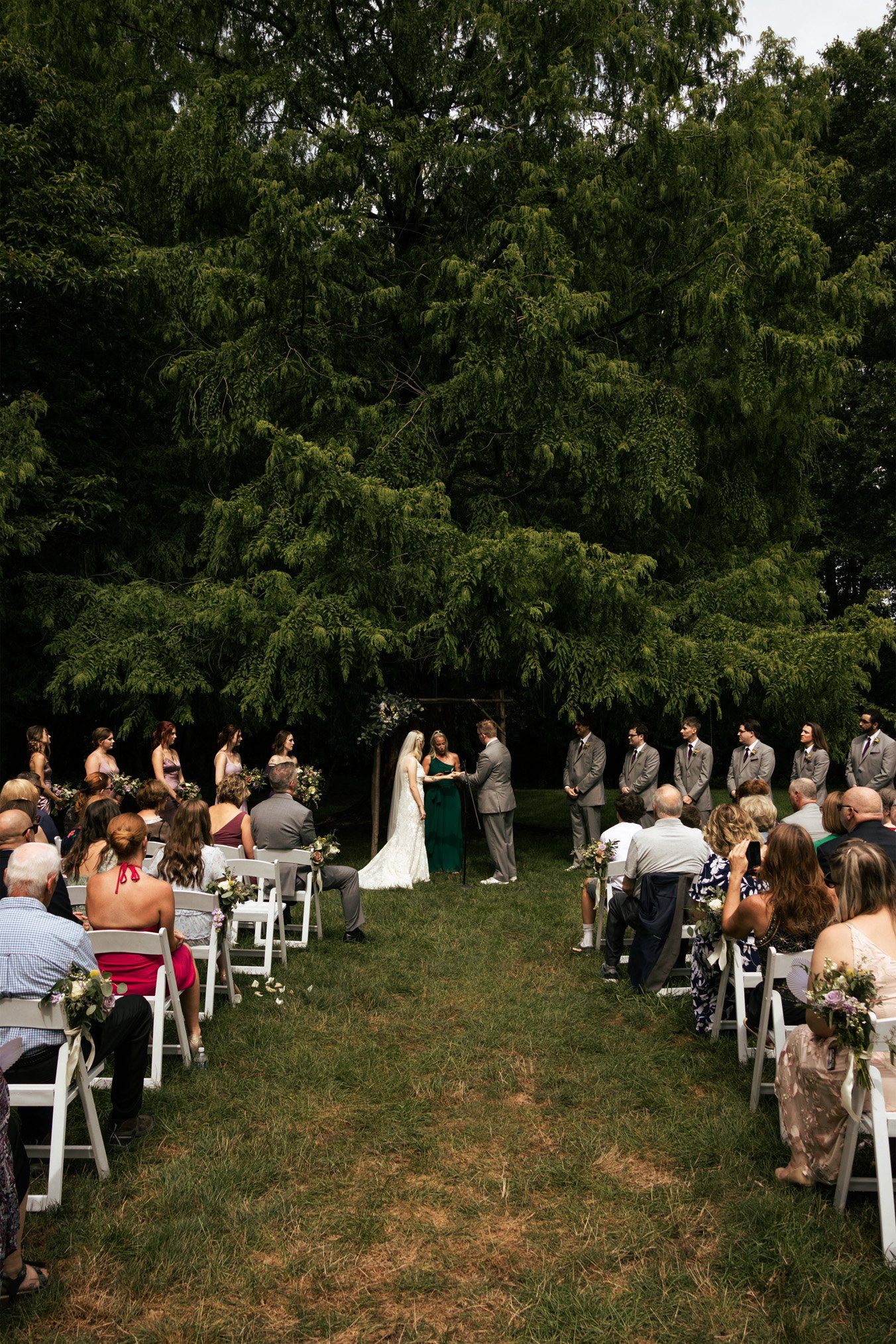 Outdoor wedding ceremony with bride and groom exchanging vows under a tree, surrounded by seated guests and bridesmaids and groomsmen in formal attire.